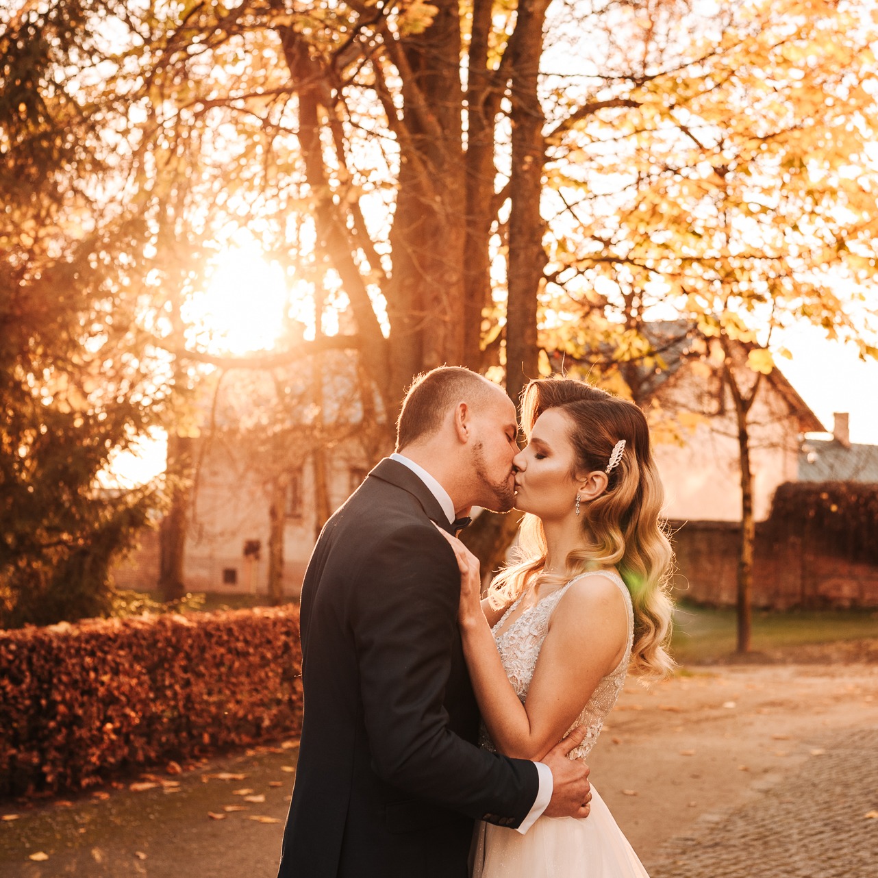 Pocałunek nowożeńców na tle jesiennego drzewa w promieniach zachodzącego słońca, panna młoda z ozdobą we włosach i subtelnym makijażem.