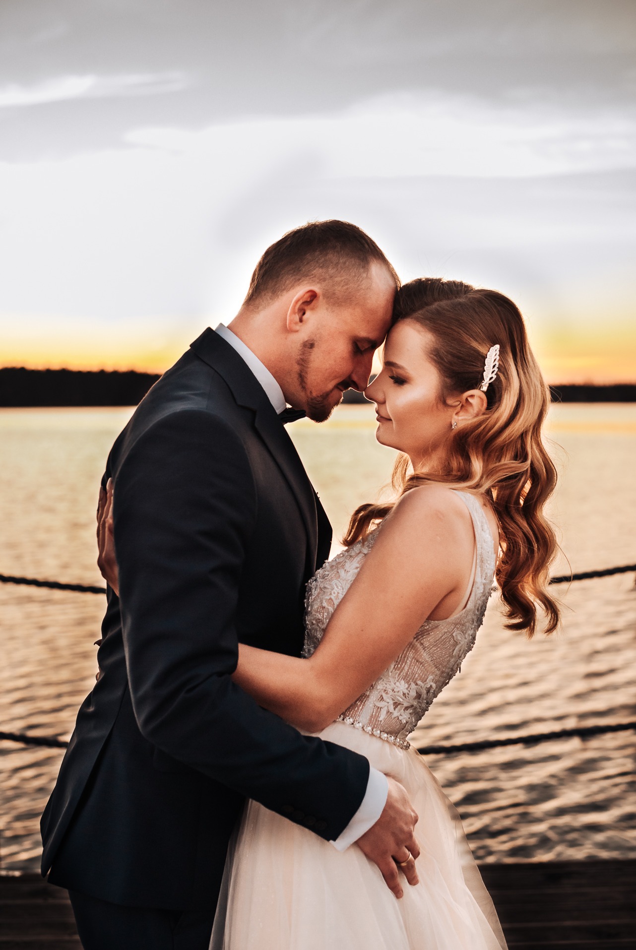 Para młoda przytulona na tle jeziora o zachodzie słońca; panna młoda ma upięcie we włosach w kształcie pióra i suknię z koronkową górą.