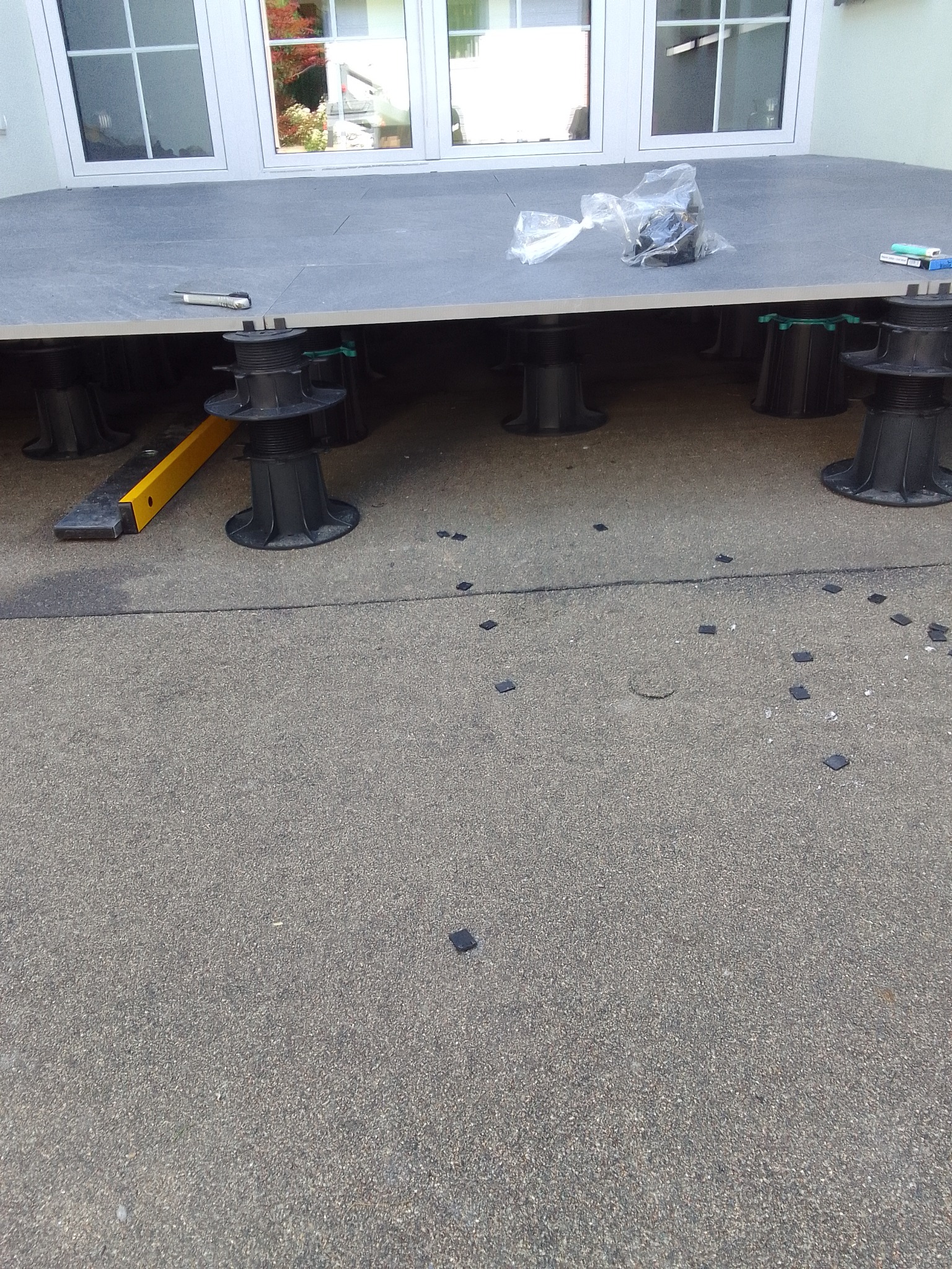 Montaż tarasu: płyty na regulowanych wspornikach, widoczne narzędzia (nóż, poziomica) i elementy mocujące, tło z oknami budynku.