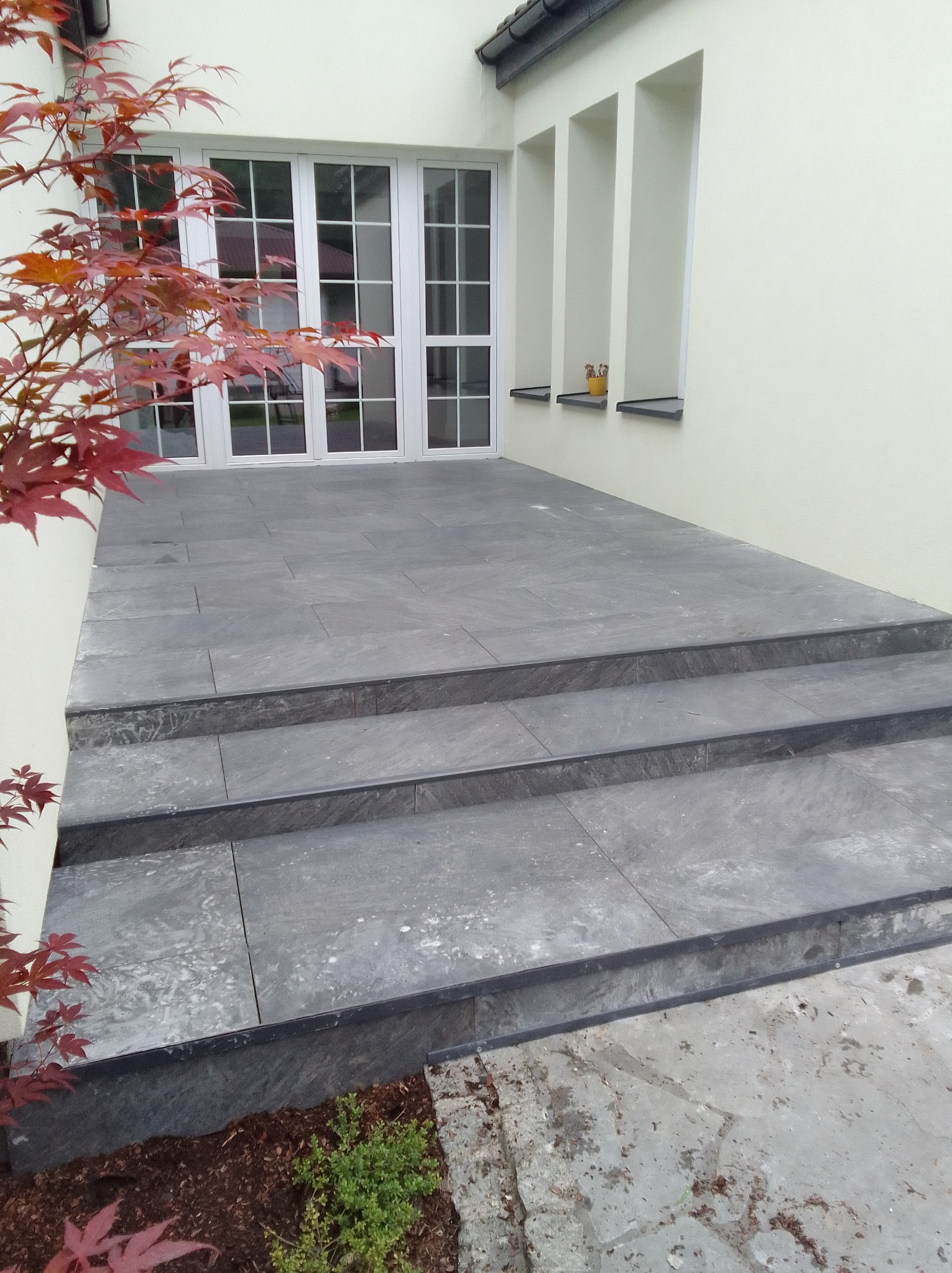 Wykończony taras z szarych, kamiennych płyt prowadzący do białych, przeszklonych drzwi balkonowych. Trzy stopnie z ciemnymi obrzeżami, obok ozdobny klon japoński i fragment rabaty. W tle kremowa...