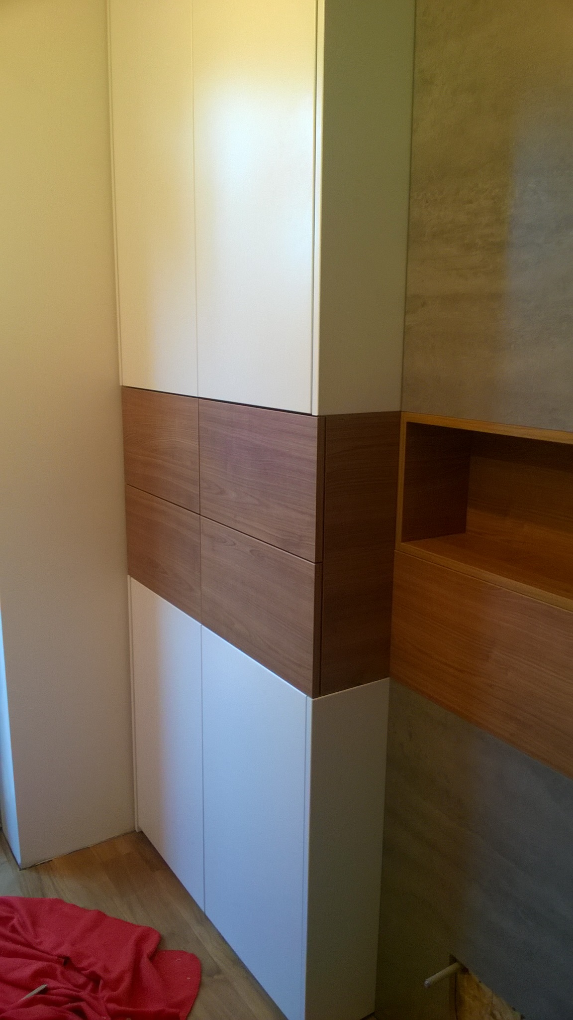 Wysoka szafa z białymi drzwiami i czterema drewnianymi szufladami, z otwartą drewnianą półką na ścianie obok, na podłodze leży czerwony materiał.