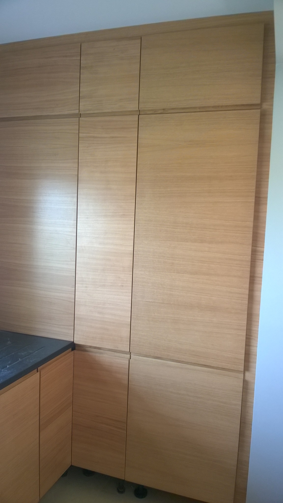 Jasne, drewniane szafki kuchenne z blatem w odcieniach szarości, tworzące minimalistyczną zabudowę pod sam sufit.