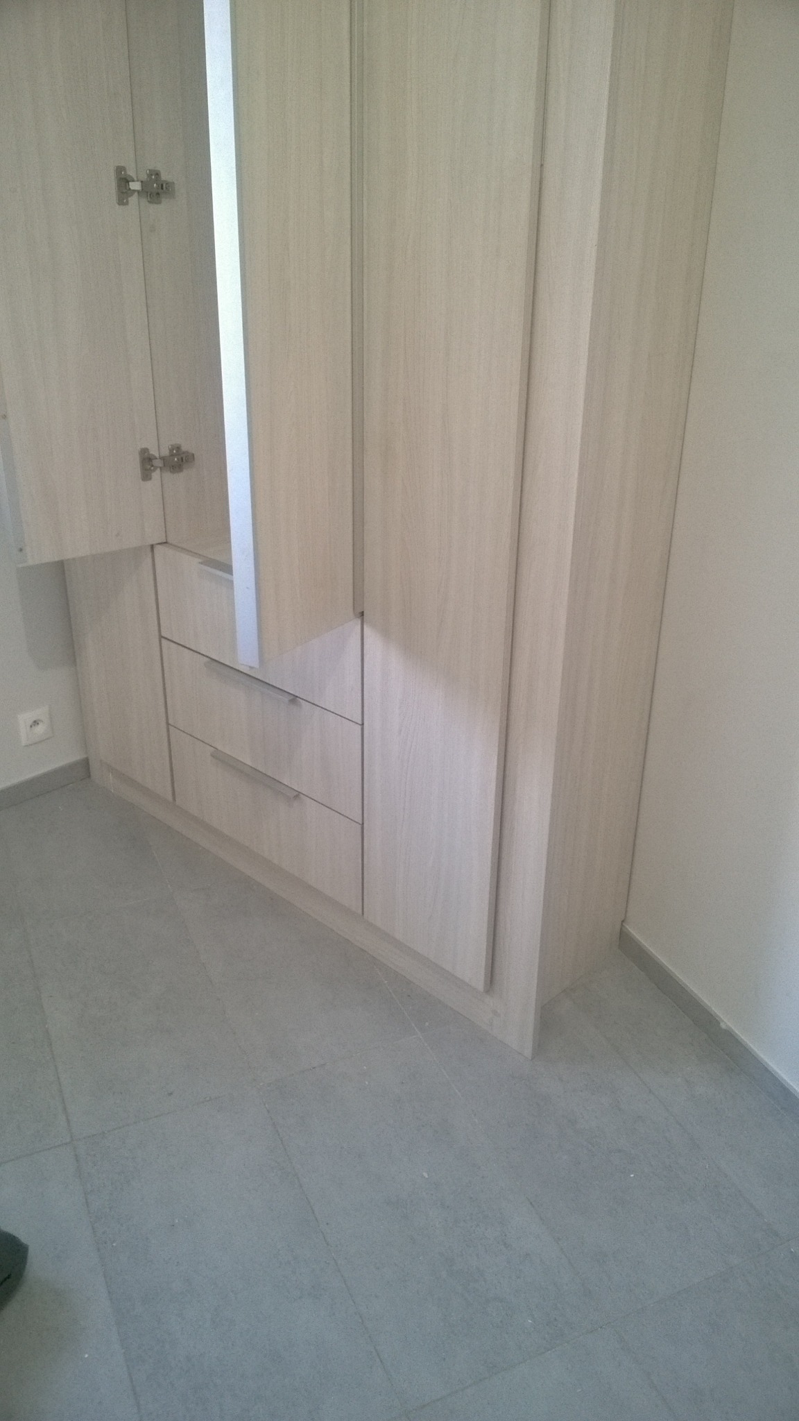 Jasna, drewnopodobna szafa z szufladami i otwartą częścią z lustrem, widoczna na tle szarej podłogi z płytek.