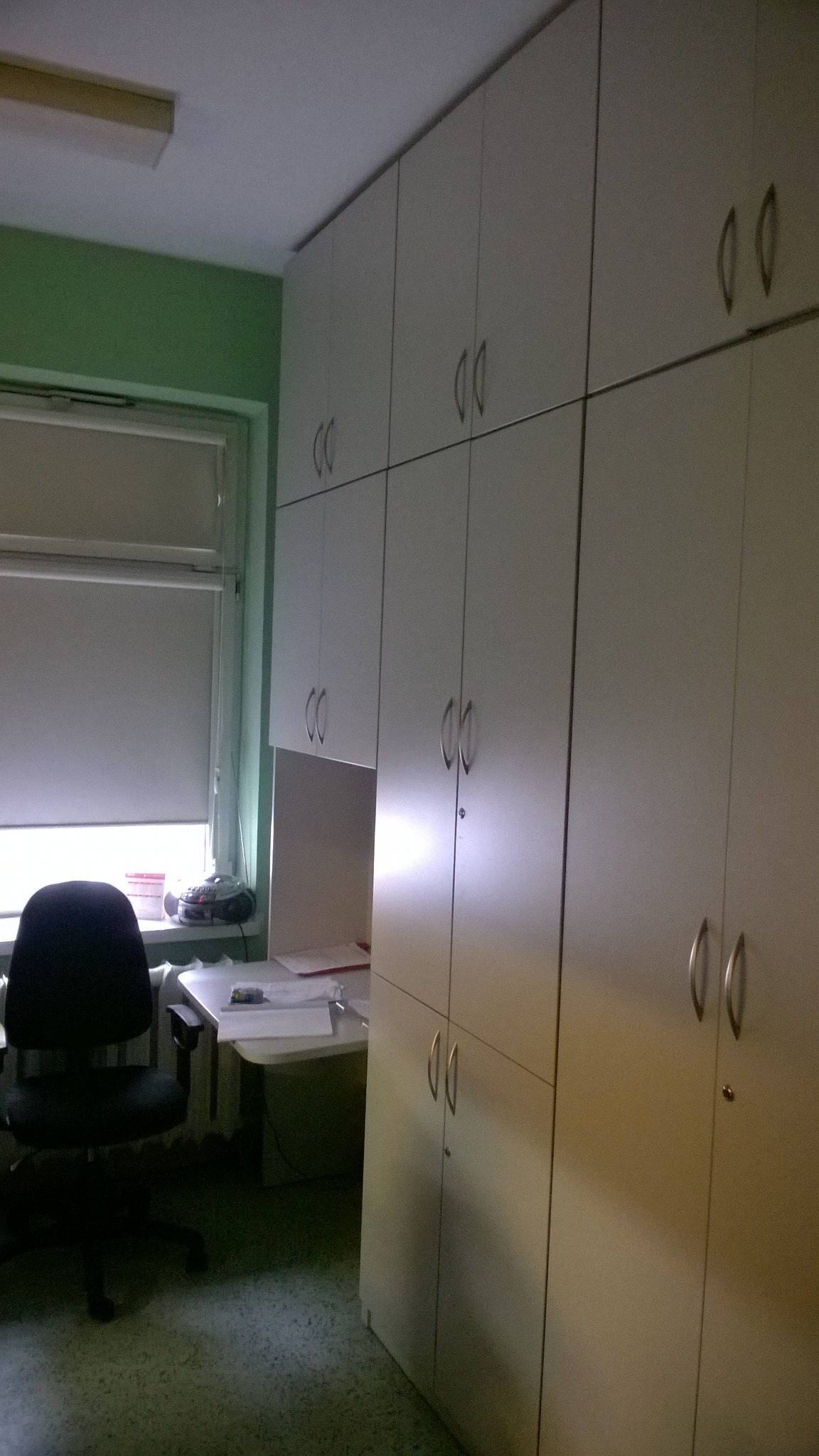 Wysoka zabudowa szaf z białymi frontami i srebrnymi uchwytami, wbudowana w pokoju z zielonymi ścianami, biurkiem i krzesłem obrotowym.