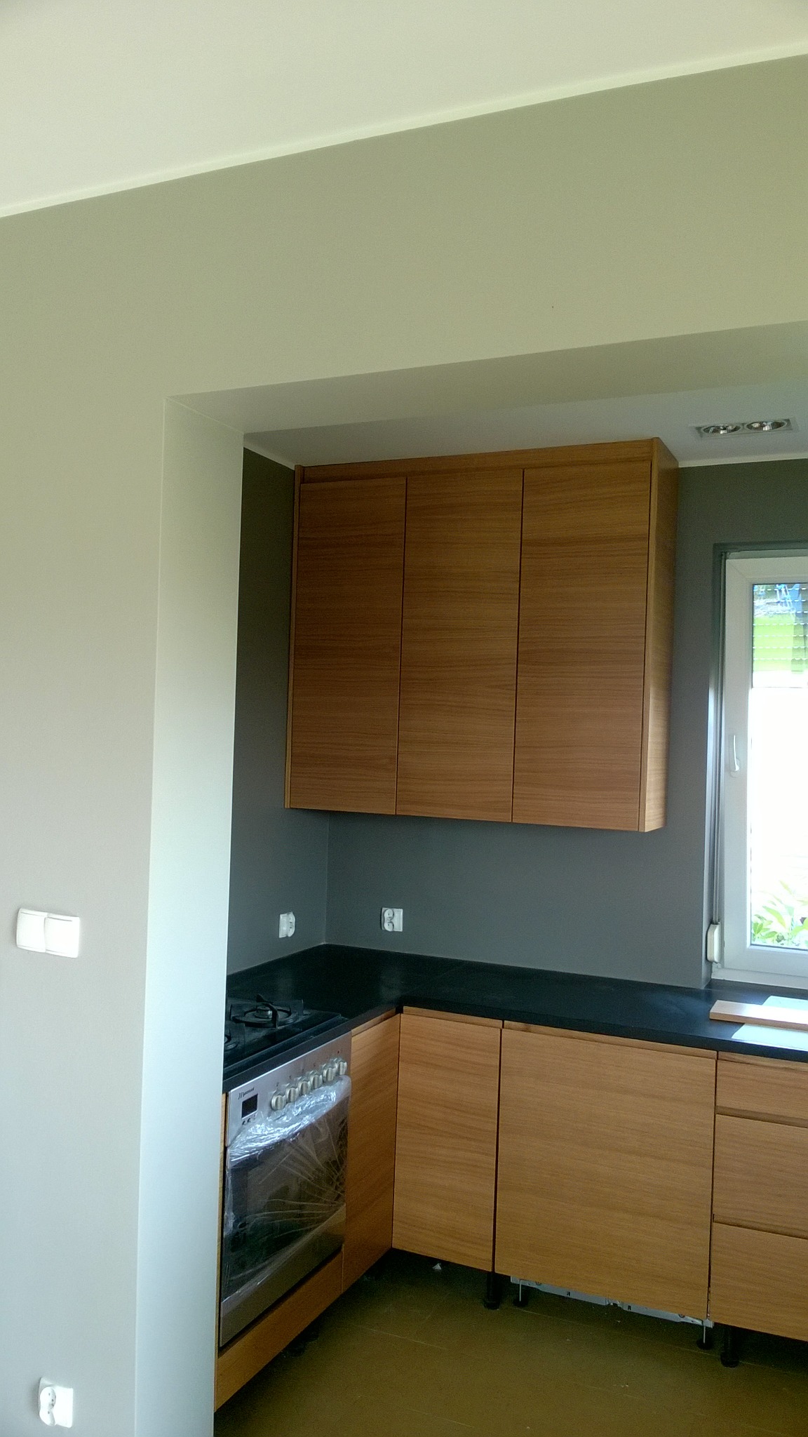 Nowoczesna kuchnia z drewnianymi frontami, czarnym blatem i piekarnikiem w folii ochronnej, widok z boku na tle szarej ściany.