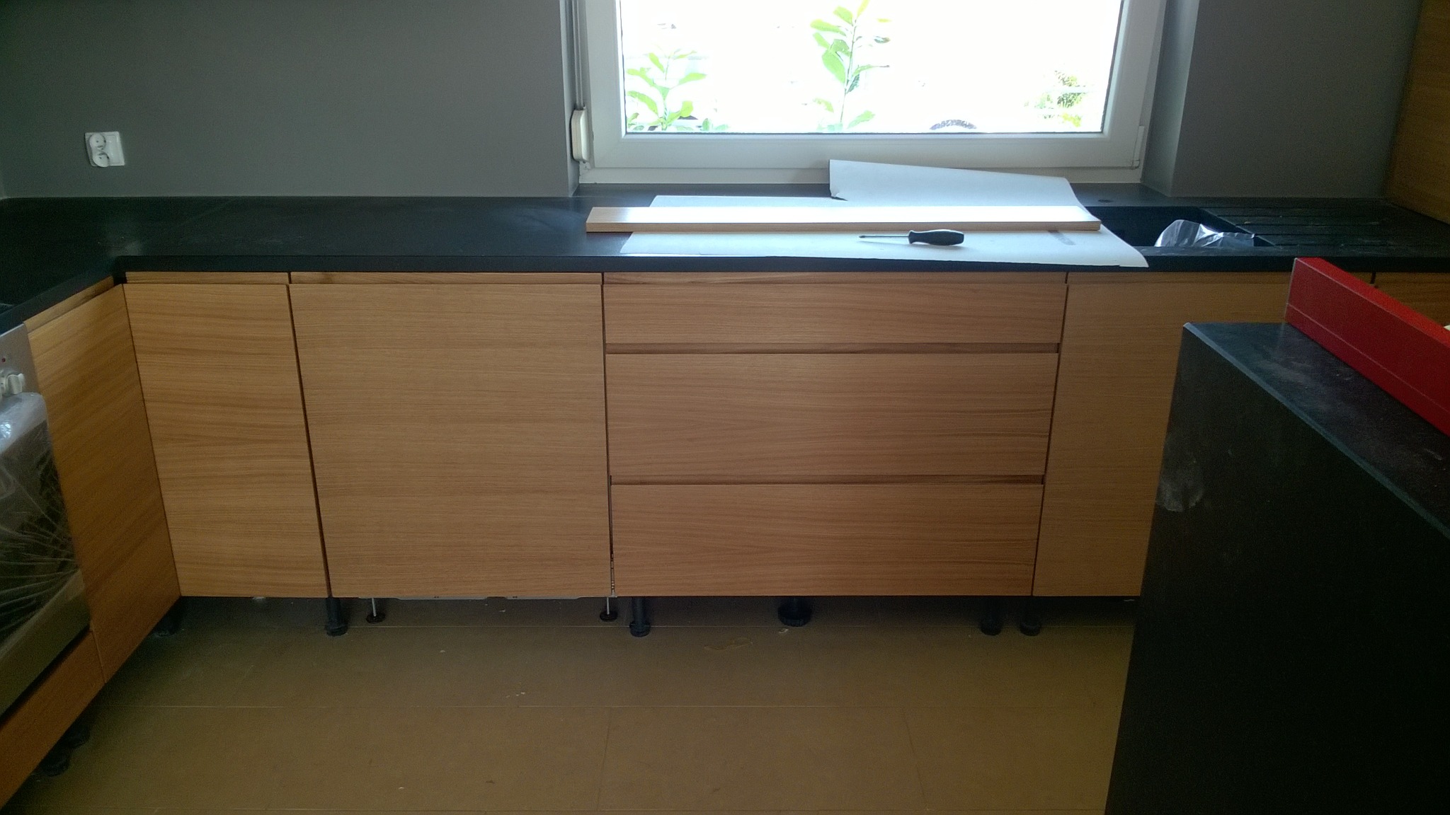 Montaż dolnych szafek kuchennych z jasnego drewna z czarnym blatem, widoczne regulowane nóżki, na blacie leżą arkusze papieru, listwa i śrubokręt.