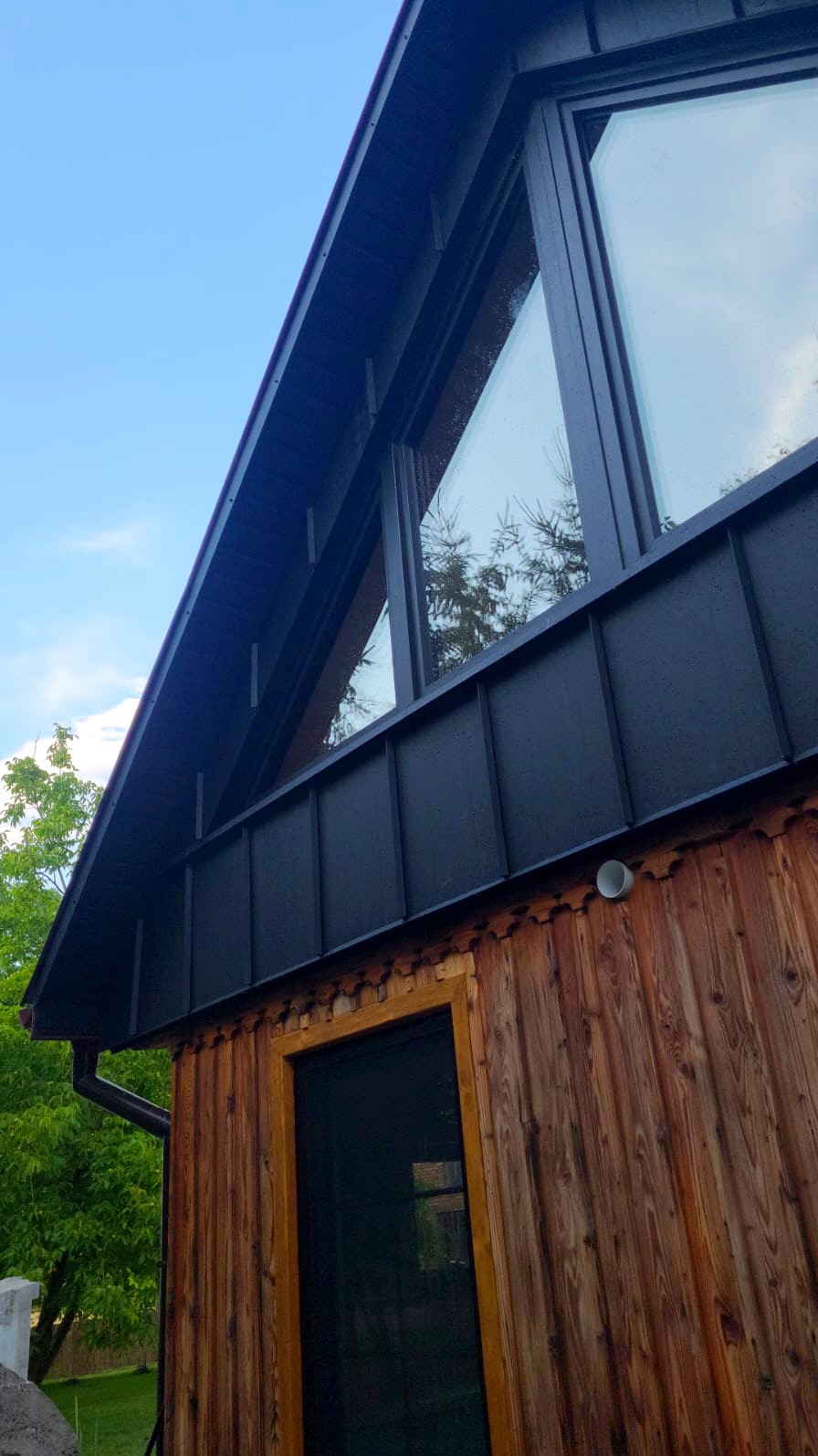 Nowoczesny dom z ciemnym dachem blaszanym i drewnianą elewacją, widok z dołu na trójkątne okno odbijające drzewa.