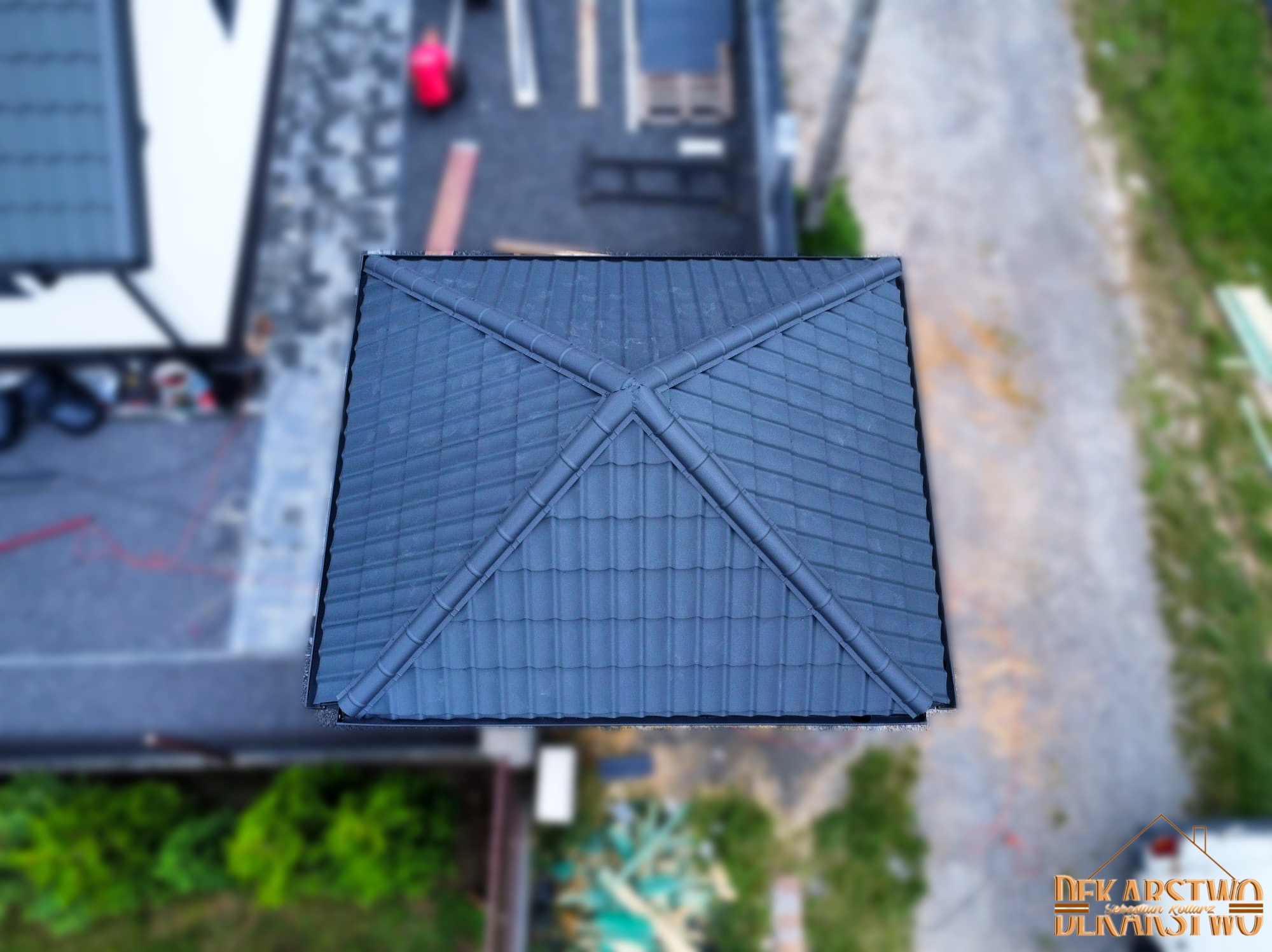 Widok z góry na nowy, ciemnoszary dach pokryty dachówką ceramiczną, z widocznymi elementami kalenicy i połaciami, na tle rozmytego otoczenia budowy i zieleni.