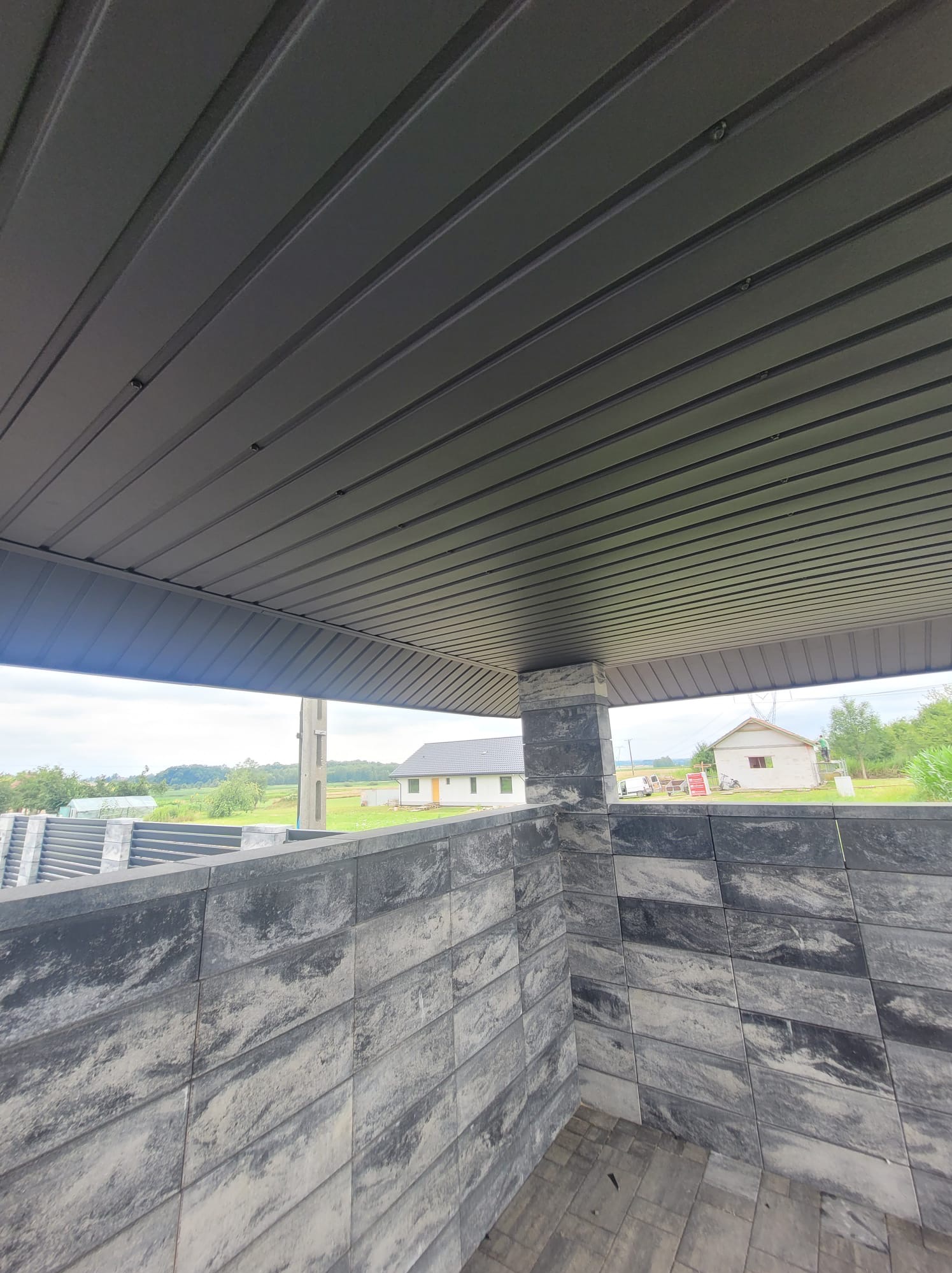 Szare, metalowe podsufitka z widocznymi śrubami mocującymi, z widokiem na krajobraz z domu i ogrodzenia z betonowych bloczków.