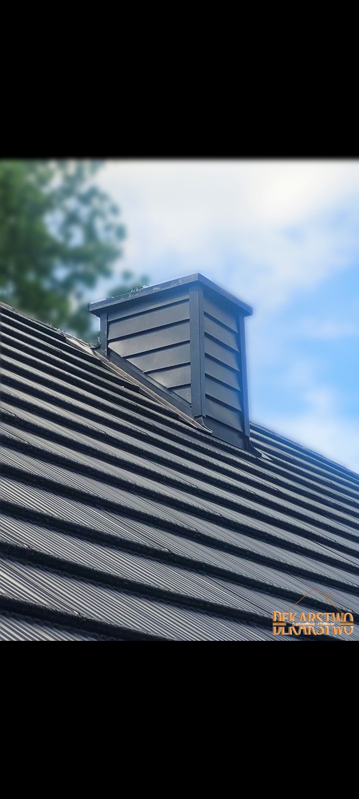 Ciemnoszary dach z blachy trapezowej z kominem w obróbce blacharskiej, widok z dołu na tle błękitnego nieba i zieleni drzew.