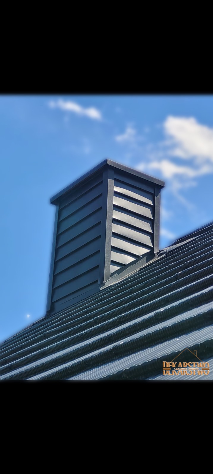 Szczegółowe ujęcie komina wentylacyjnego z czarnymi, poziomymi szczelinami na dachu z blachy trapezowej, na tle błękitnego nieba z delikatnymi obłokami.