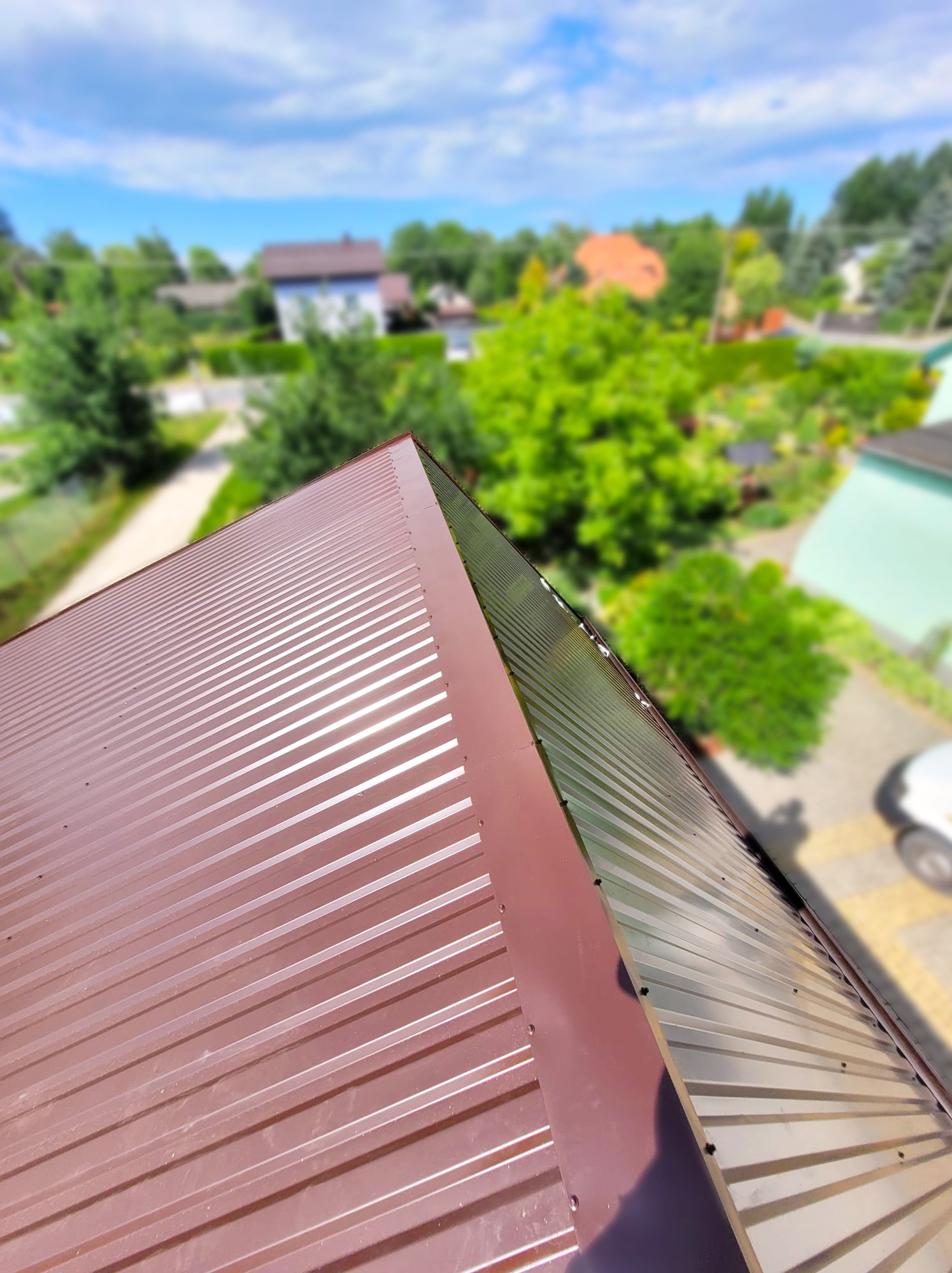 Nowe, brązowe pokrycie dachowe z blachy trapezowej widziane z góry, z widokiem na okoliczne domy i zieleń w tle.