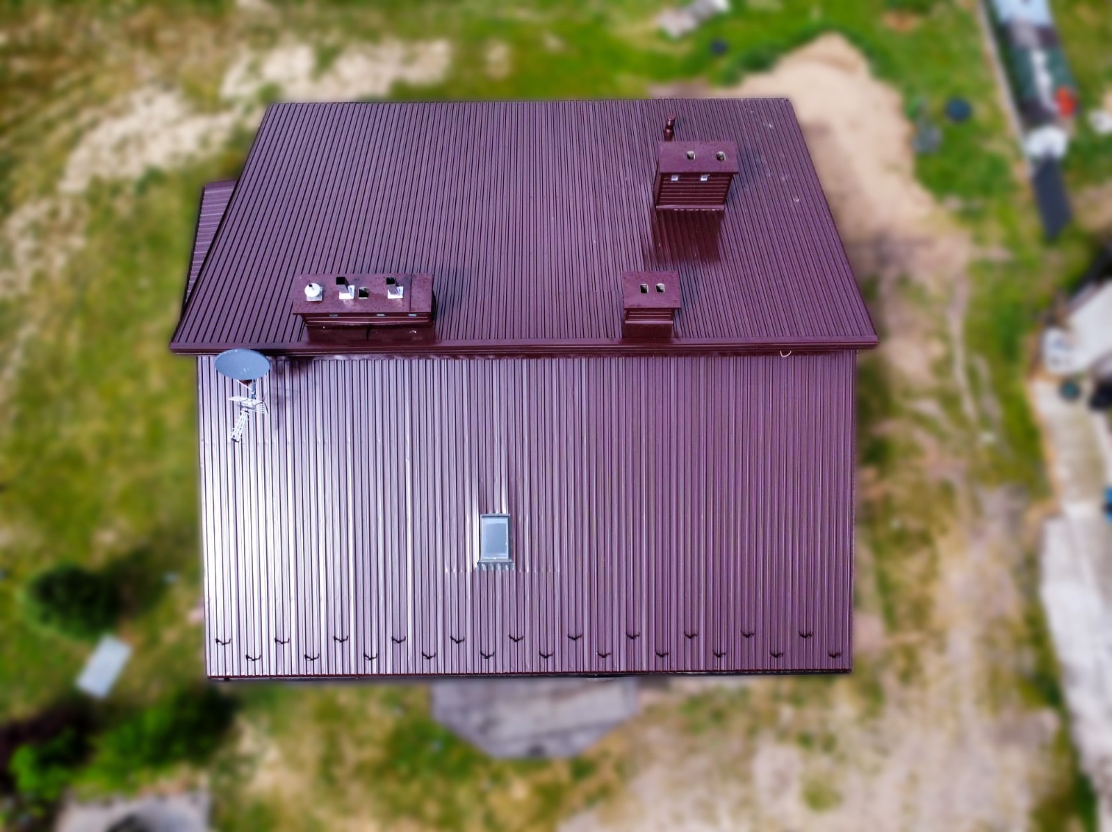 Widok z góry na dom z bordowym, metalowym dachem z blachy trapezowej, widoczne kominy, okno dachowe i antena satelitarna, w tle zamazane otoczenie zieleni.