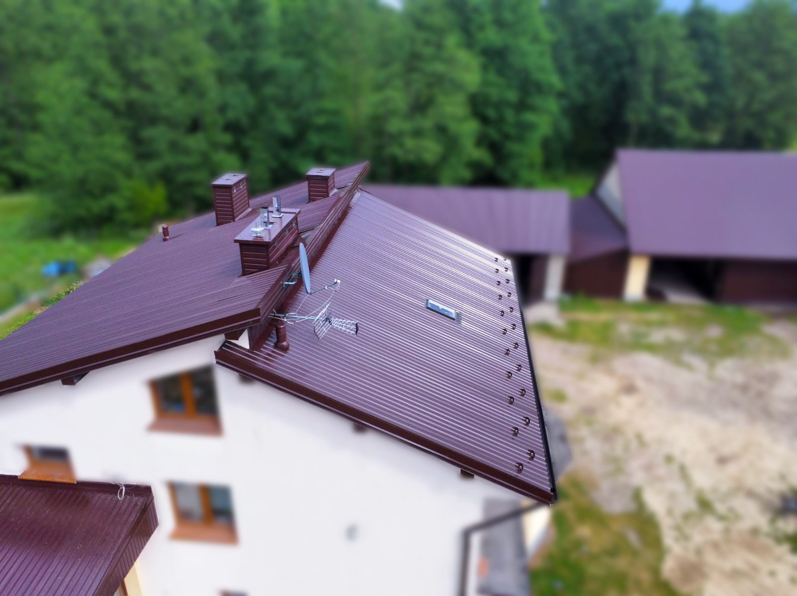 Widok z góry na dom z nowym, bordowym dachem z blachy trapezowej, widoczna antena satelitarna, kominy i okno dachowe, w tle zieleń drzew.