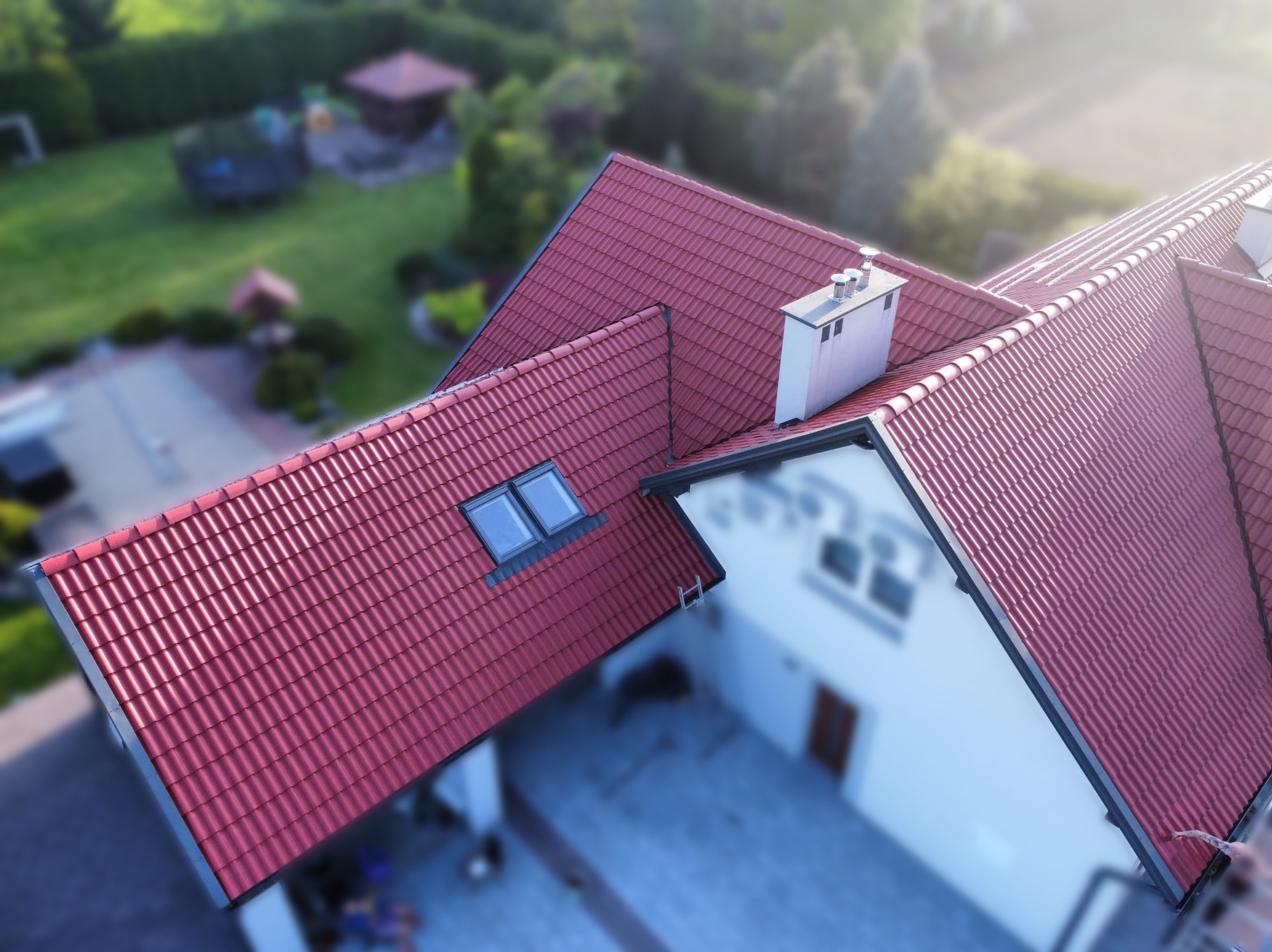 Nowy, czerwony dach z dachówki ceramicznej z oknami dachowymi i kominem na jasnym domu z zadbanym ogrodem, sfotografowany z góry.