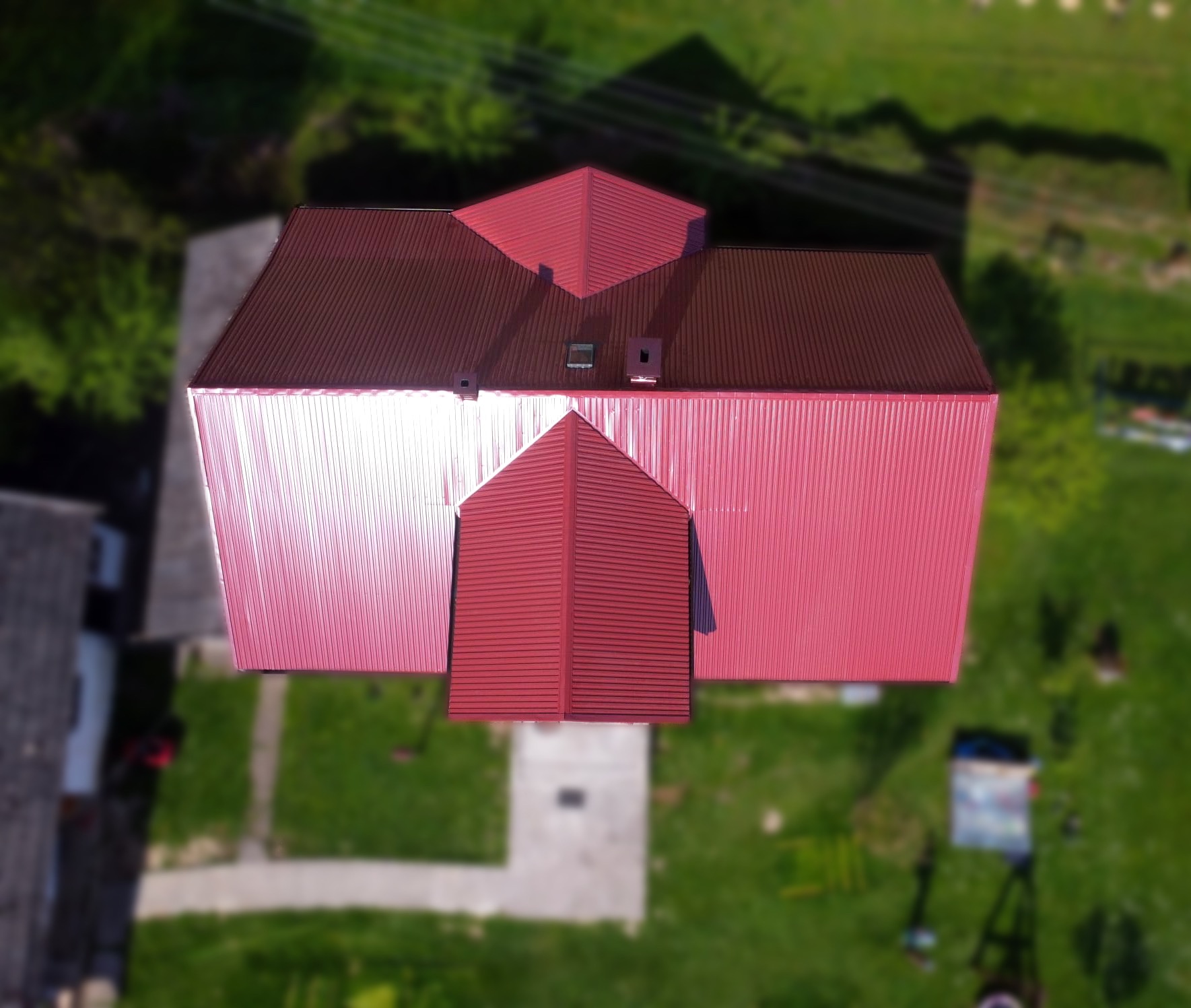 Widok z lotu ptaka na dom z nowym, czerwonym dachem z blachy trapezowej, z widocznymi kominami wentylacyjnymi i elementami obróbek blacharskich. W tle rozmyty krajobraz zieleni.