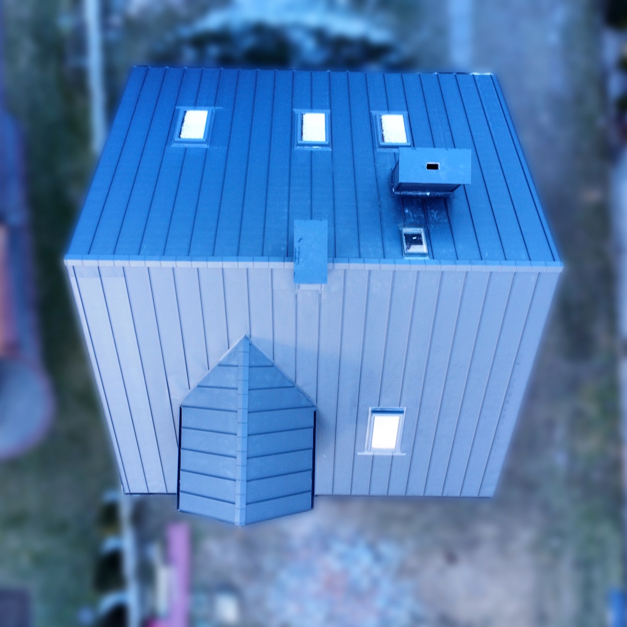 Widok z góry na dom z blachy trapezowej w odcieniach szarości i błękitu, z lukarnami, kominem i małą dobudówką nad wejściem. Blacha ułożona pionowo na ścianach i dachu.