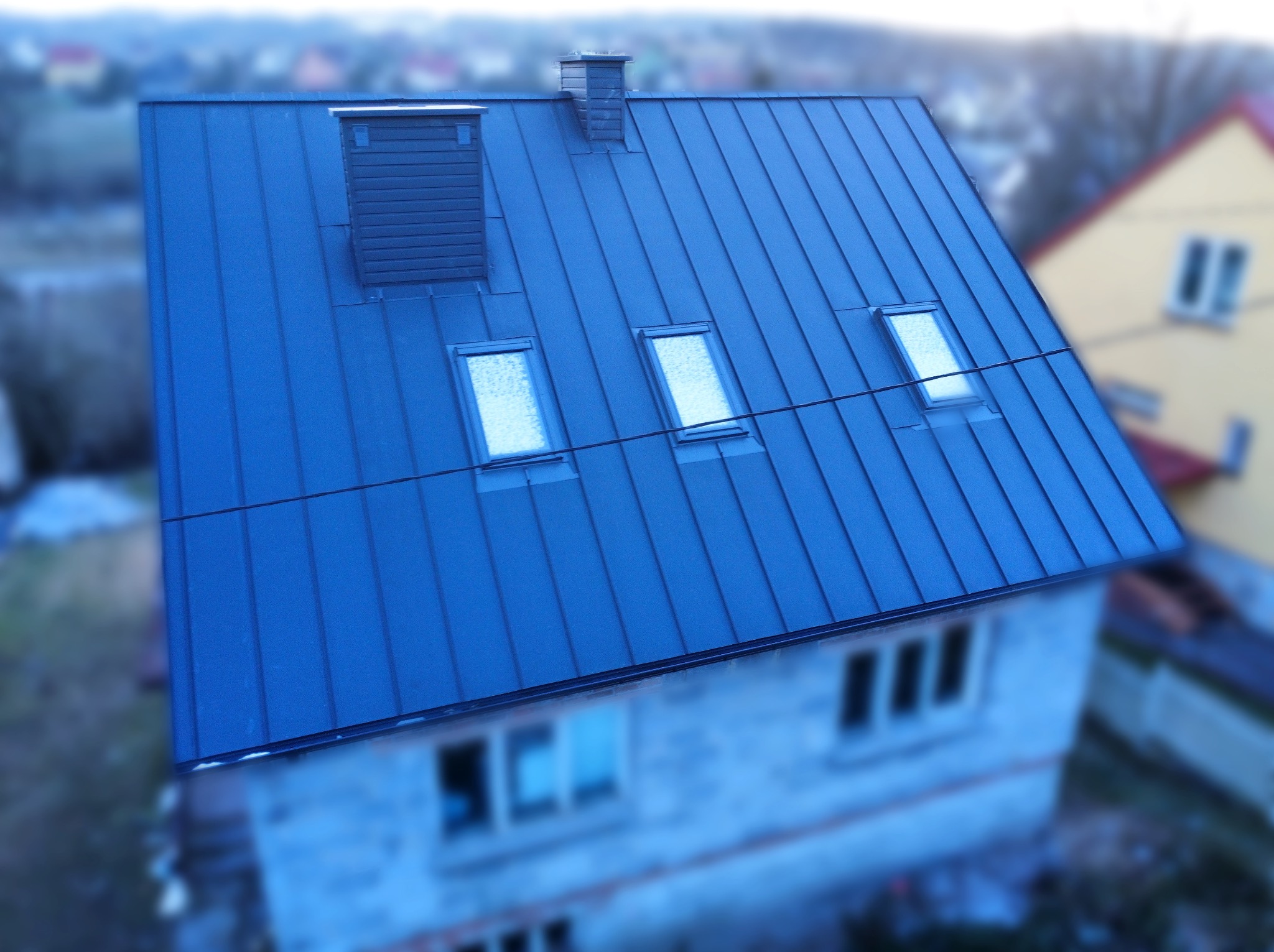 Niebieski dach z blachy na domu z trzema oknami dachowymi i dwoma kominami, widziany z góry pod kątem.