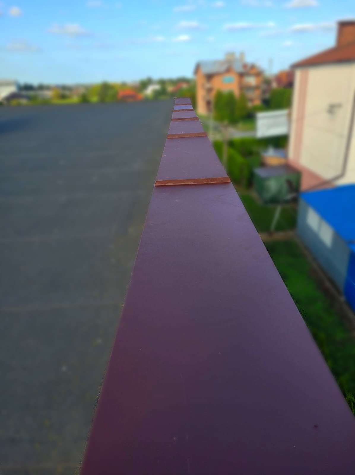 Widok z góry na fragment dachu z blachy w kolorze burgundowym, z widocznymi elementami łączenia i tłem rozmytego krajobrazu miejskiego.