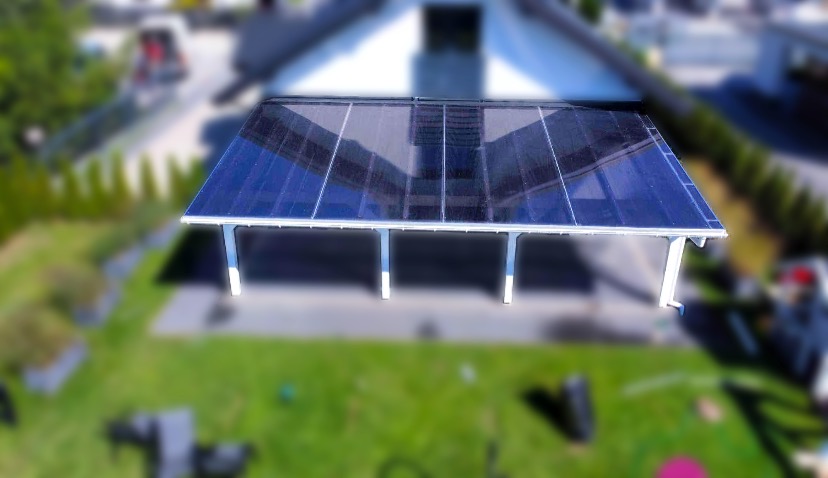 Widok z góry na zadaszenie tarasu z przezroczystego poliwęglanu, wsparte na białych słupach, z widocznym ogrodem i budynkiem w tle, z efektem bokeh.