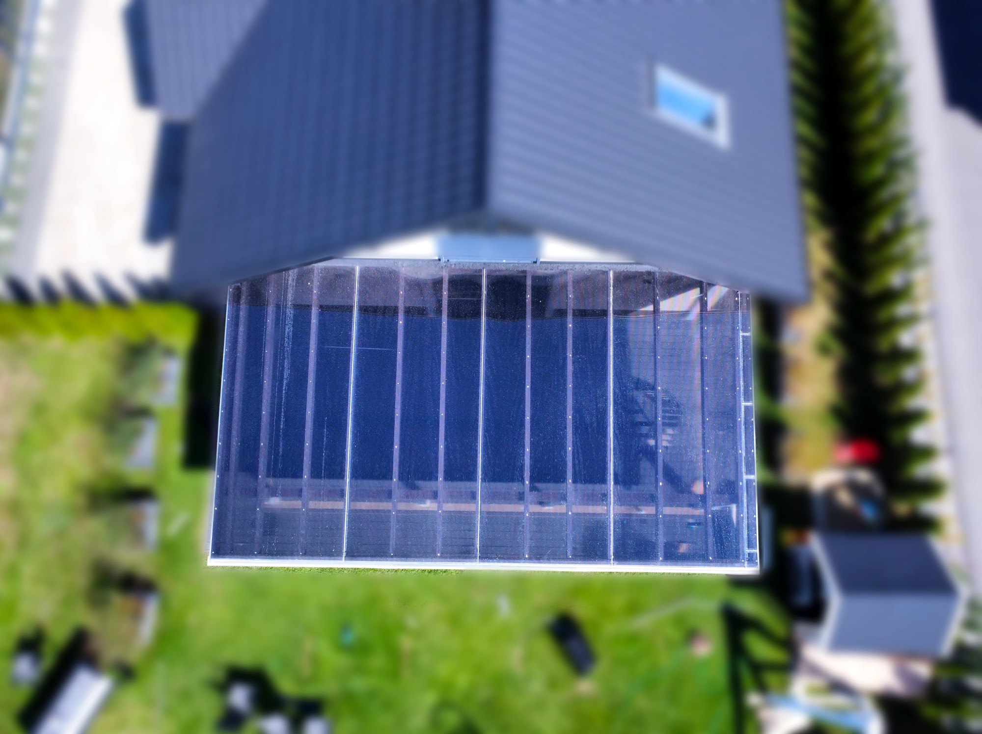 Widok z góry na przezroczysty dach z poliwęglanu nad tarasem, przylegający do nowoczesnego, grafitowego domu z oknem dachowym; rozmyte tło zielonego trawnika.