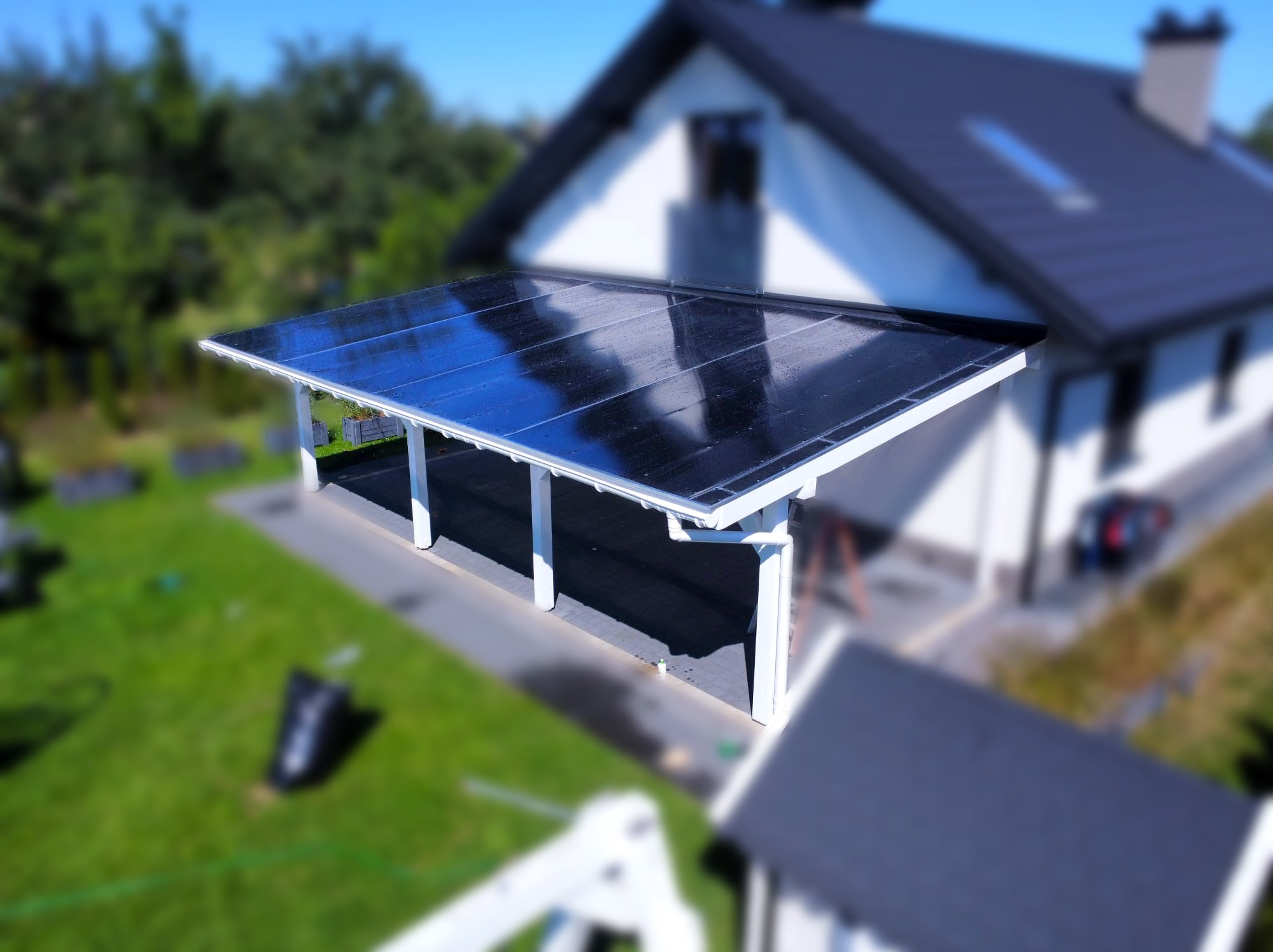 Nowoczesna wiata samochodowa z przezroczystym, ciemnym zadaszeniem, wsparta na białych kolumnach, na tle domu z ciemnym dachem i zieleni. Widok z góry.