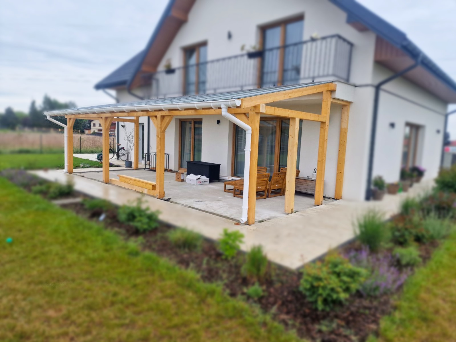 Drewniana pergola z przezroczystym zadaszeniem przy białym domu w Żabnie. Widoczne rynny, meble ogrodowe i zadbany trawnik. Ujęcie z poziomu gruntu.