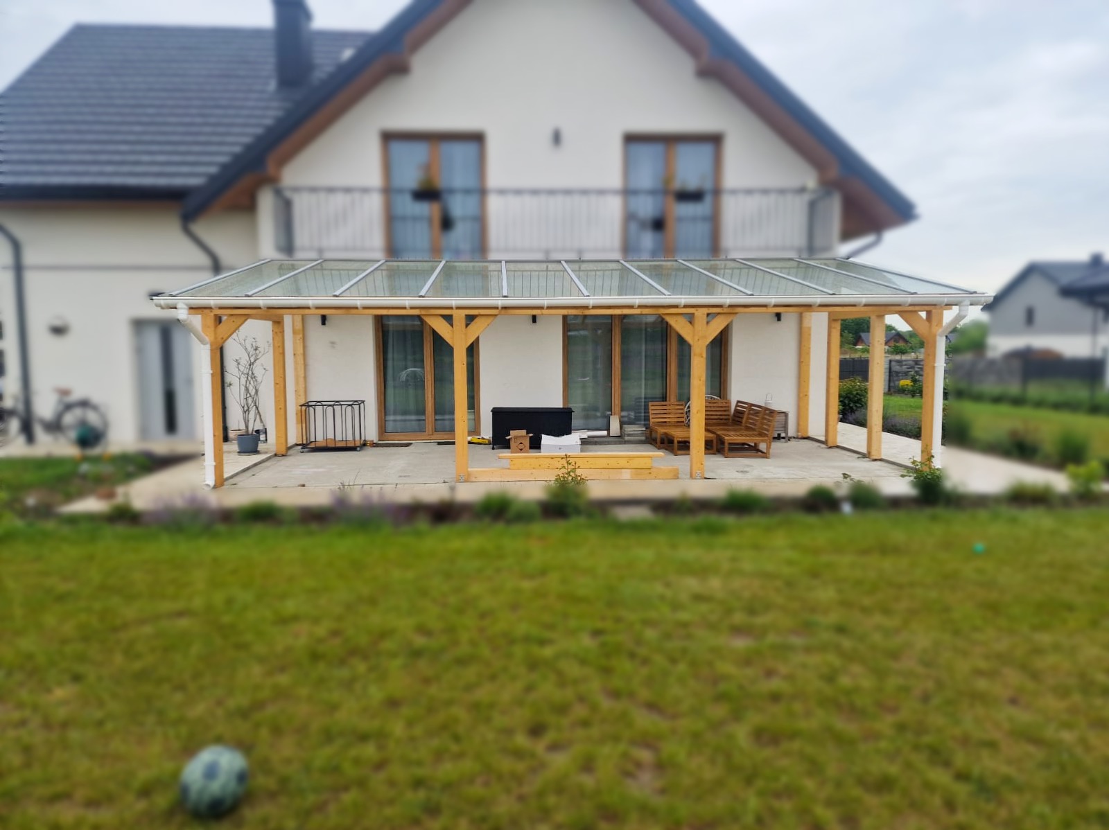 Zadaszenie tarasu z drewnianą konstrukcją i przezroczystym dachem, widok z ogrodu. Na tarasie meble ogrodowe i elementy w trakcie montażu. Dom w tle.