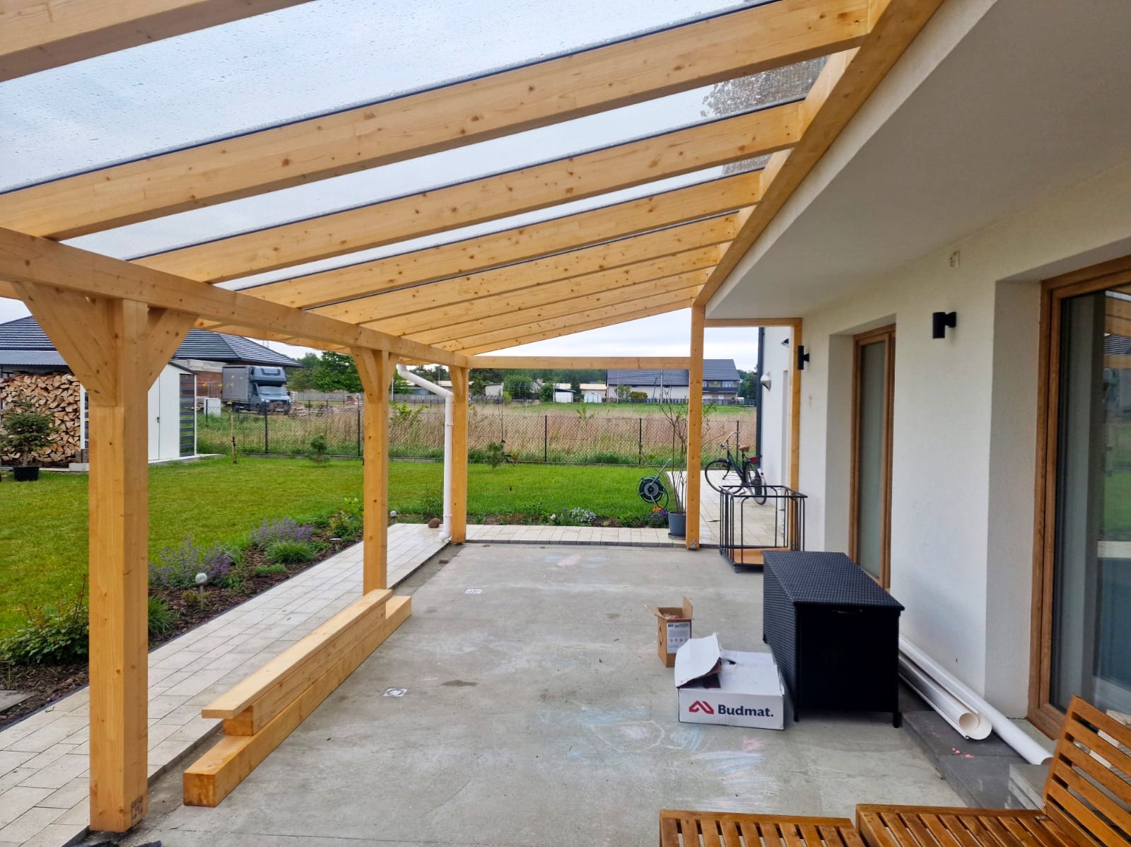 Drewniana pergola z przezroczystym zadaszeniem przylegająca do białego budynku. Widoczne elementy konstrukcyjne, ogród i pudełko Budmat na betonowej posadzce.
