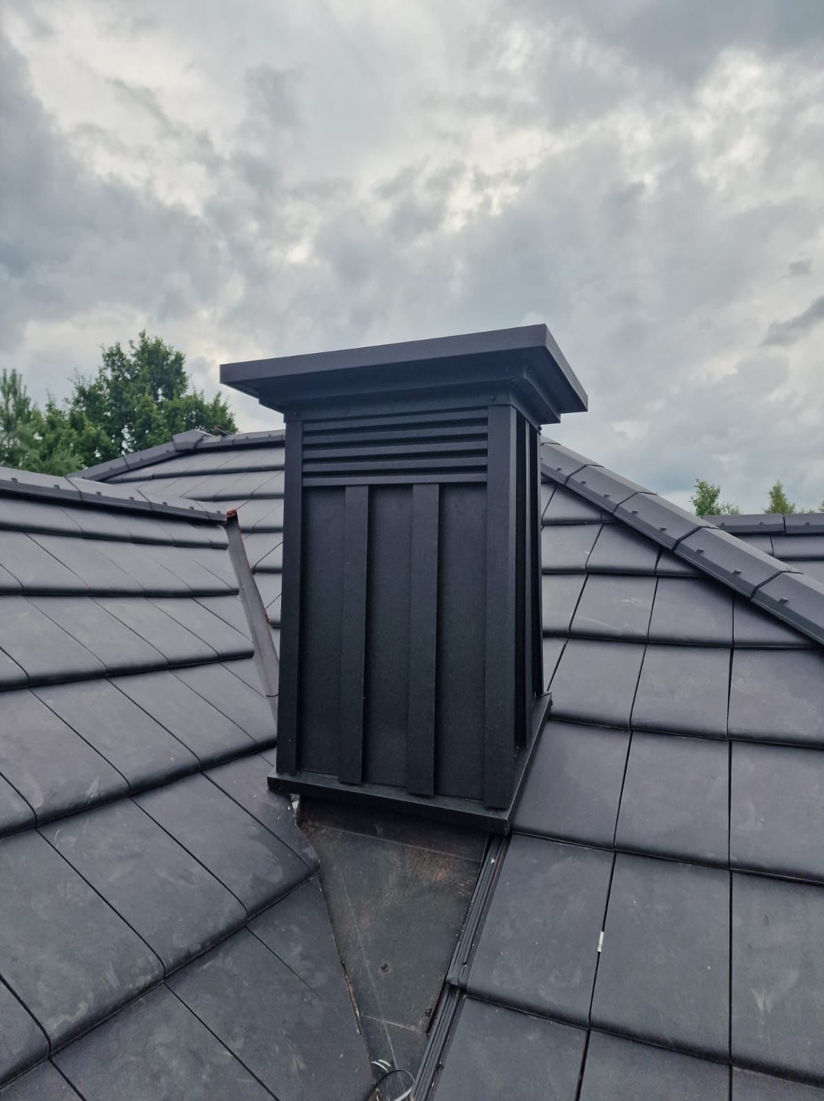 Czarny komin wentylacyjny na dachu z ciemnej dachówki ceramicznej. Widok z dołu, pochmurne niebo w tle. Komin z poziomymi szczelinami wentylacyjnymi.