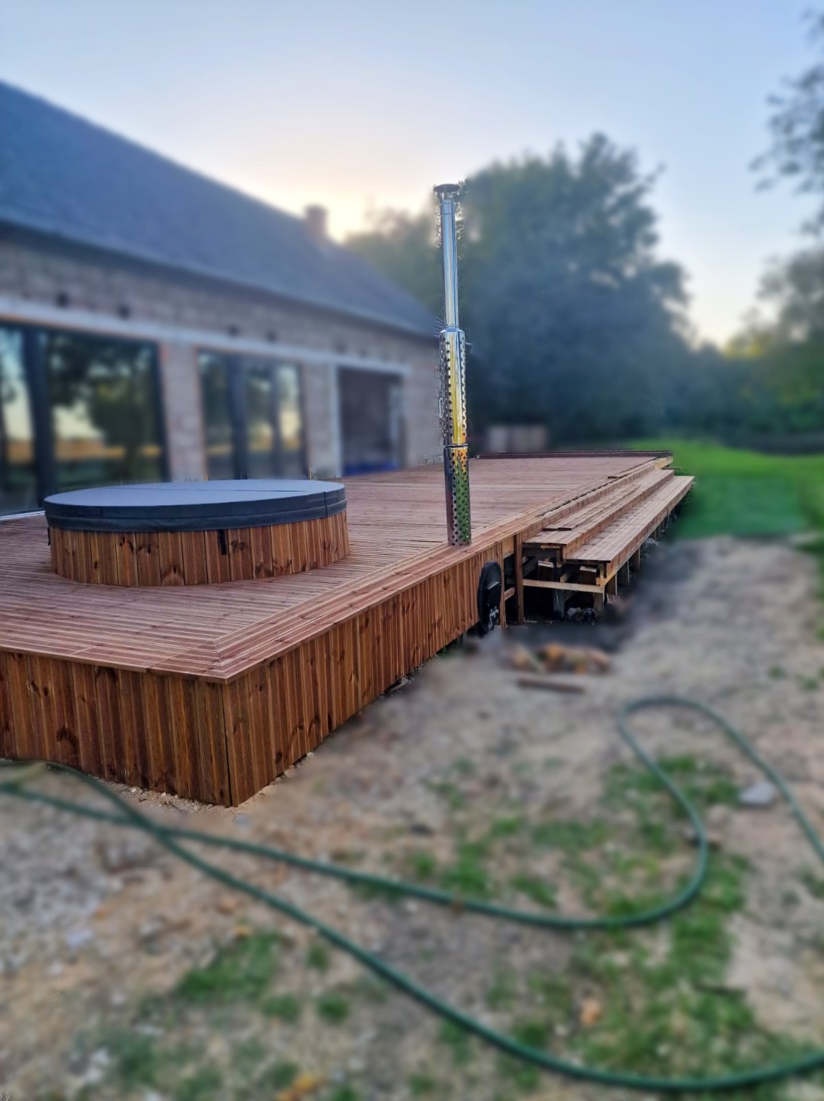 Drewniany taras z jacuzzi i kominem na tle domu z dużymi oknami. Taras wykonany z desek w ciepłym odcieniu brązu, otoczony zielenią. Widok na konstrukcję tarasu.