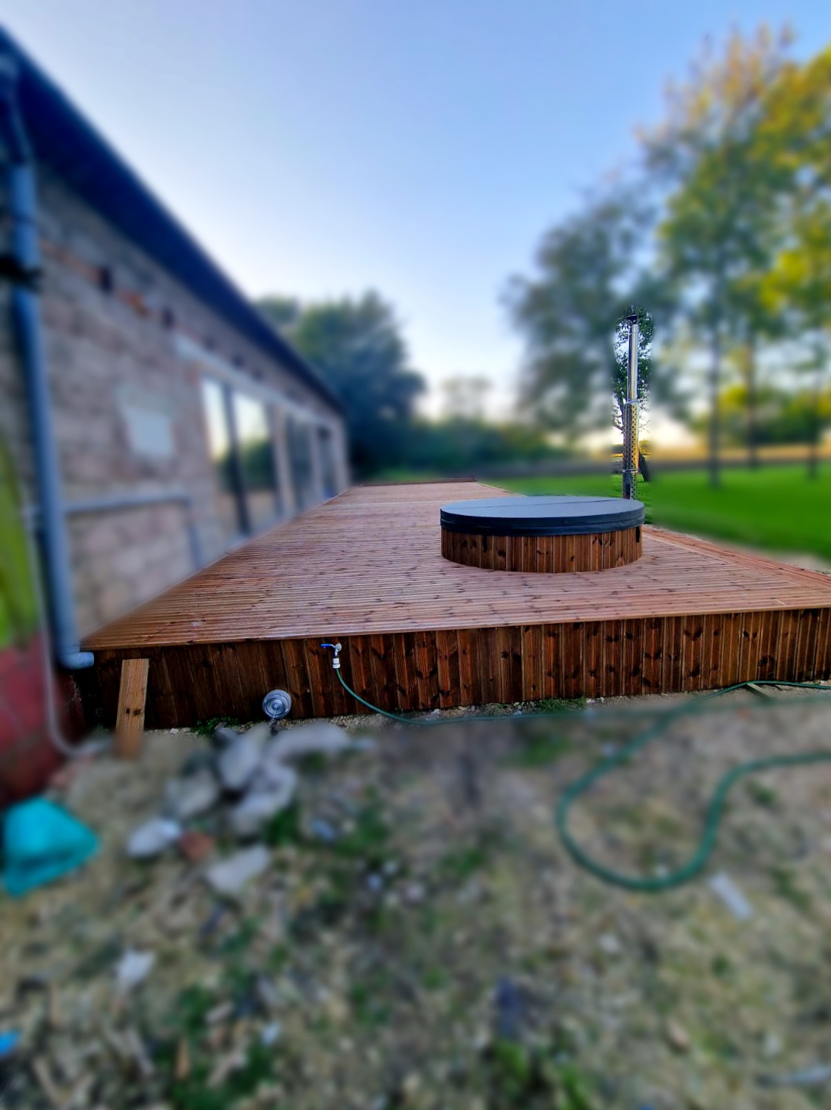 Drewniany taras z jacuzzi na zewnątrz, widok z dołu. Widoczne elementy konstrukcyjne, złącza i instalacja wodna. Tło stanowi budynek i zieleń.