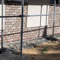 Montaż brązowych płytek klinkierowych na elewacji budynku, widoczne rusztowanie i elementy mocujące płytki.