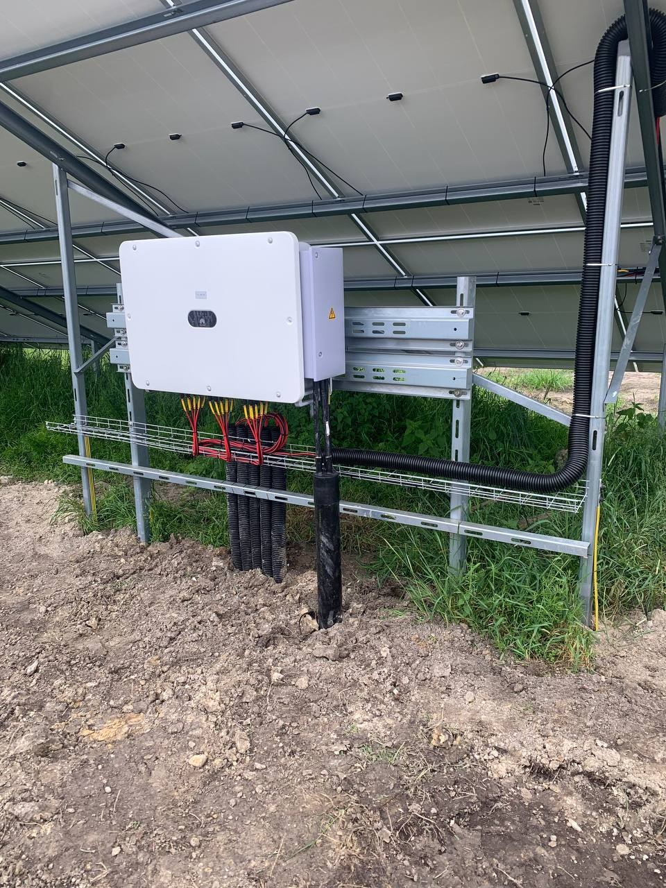 Widok od spodu na fragment konstrukcji farmy fotowoltaicznej z zamontowanym inwerterem w białej obudowie, widoczne kable w czarnych peszlach oraz uziemienie, trawa i ziemia w tle.
