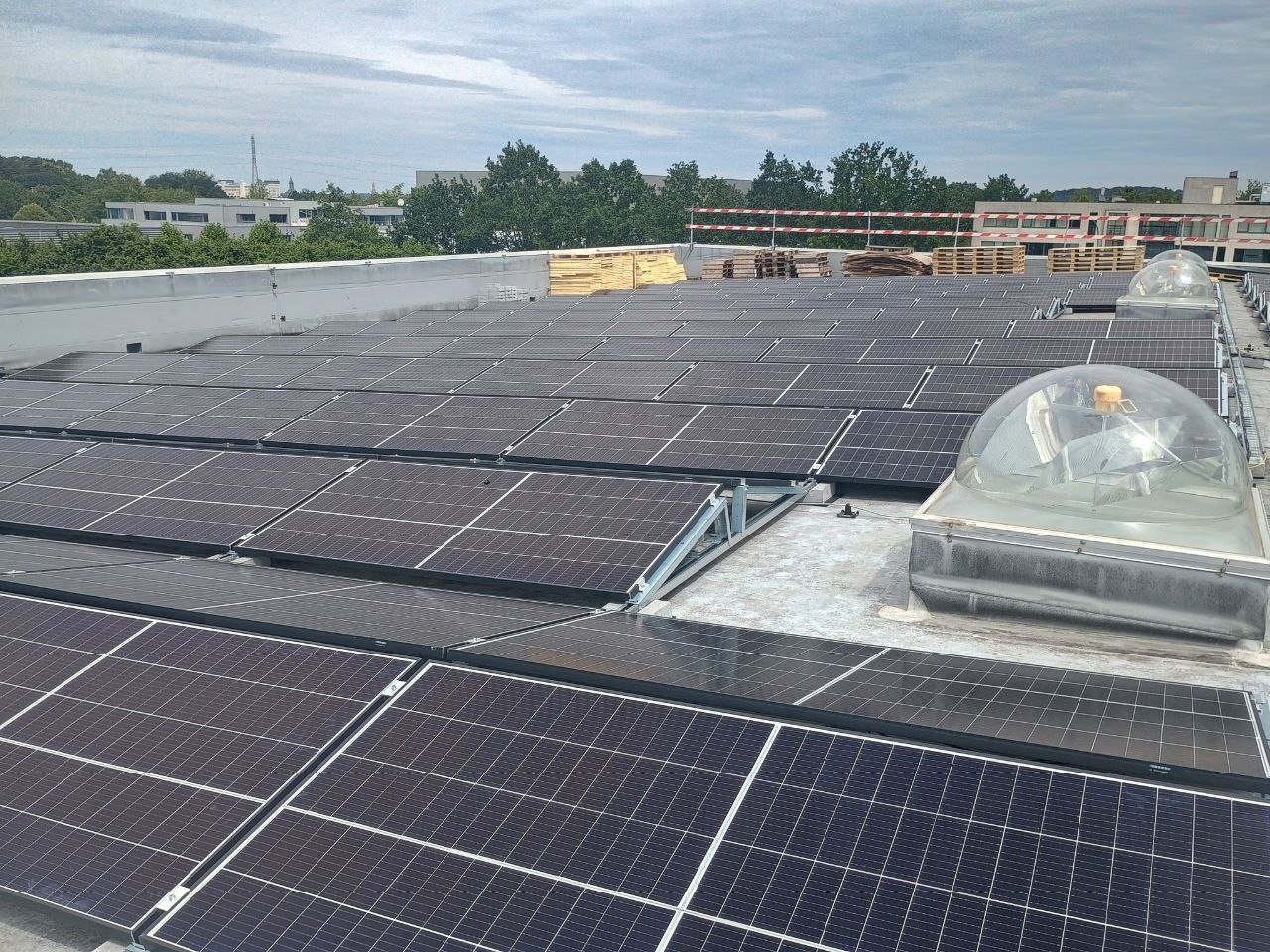 Widok z góry na panele fotowoltaiczne zamontowane na płaskim dachu budynku przemysłowego, z widocznymi świetlikami kopułowymi i paletami z materiałami budowlanymi w tle.