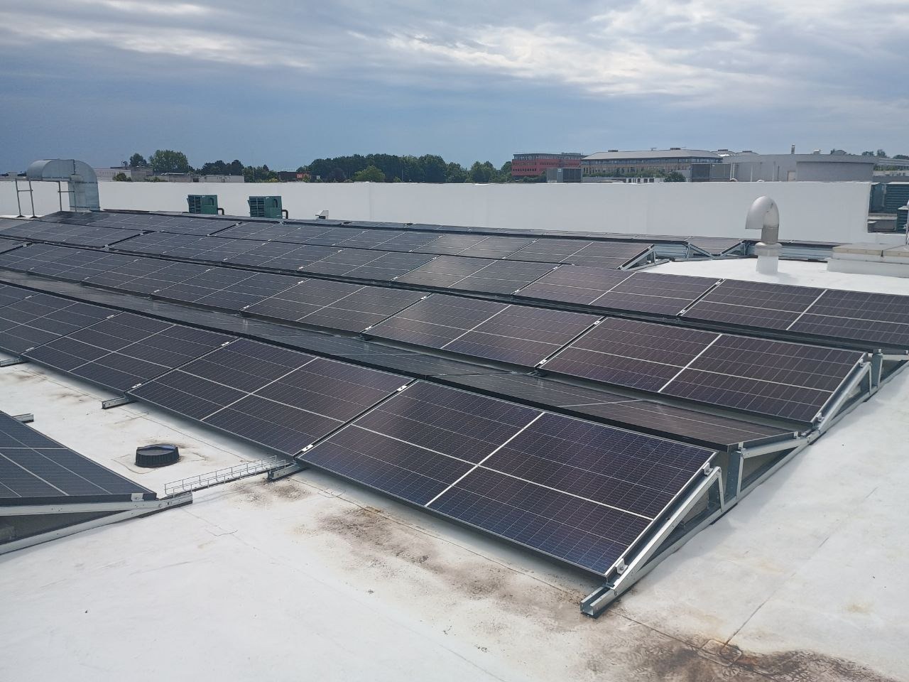 Rząd paneli fotowoltaicznych na płaskim, lekko zabrudzonym dachu, z widoczną konstrukcją wsporczą i elementami wentylacji w tle, pod pochmurnym niebem.