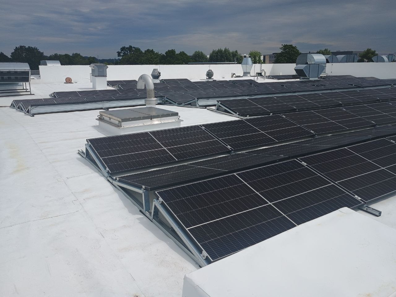Instalacja paneli słonecznych na płaskim, białym dachu budynku komercyjnego; widoczne liczne panele w rzędach, metalowe konstrukcje wsporcze, system wentylacyjny i pochmurne niebo w tle.