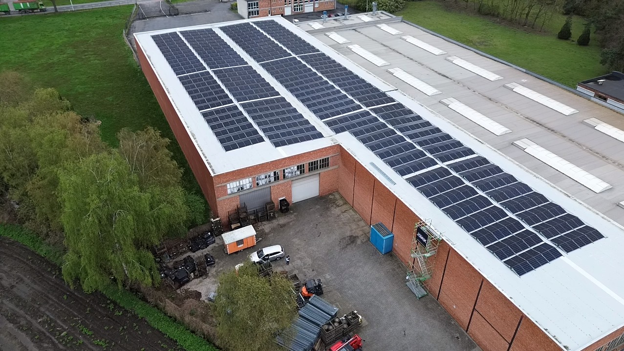 Widok z góry na duży budynek przemysłowy z zamontowanymi panelami fotowoltaicznymi na dachu, obok budynku zaparkowane pojazdy i kontenery.