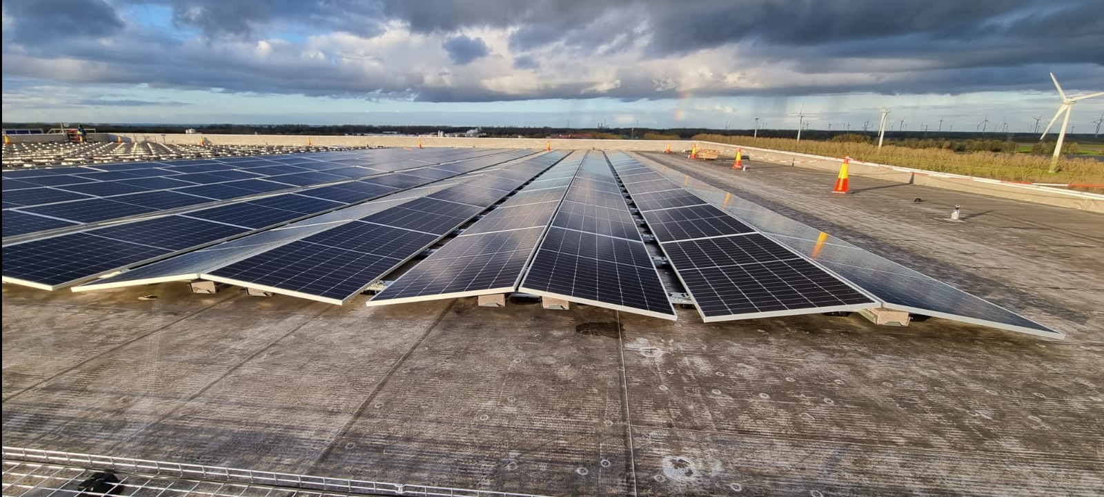 Rząd paneli fotowoltaicznych na płaskim dachu przemysłowym w pochmurny dzień, w tle widoczne turbiny wiatrowe i tęcza.