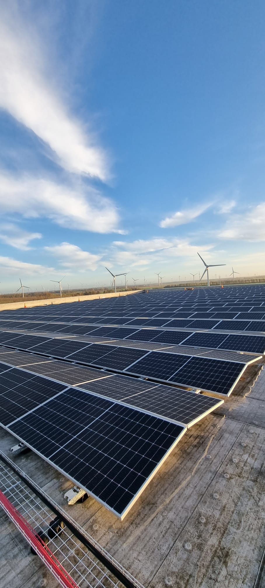 Rozległa instalacja fotowoltaiczna na dachu, z licznymi panelami ustawionymi w rzędach, a w tle widoczne turbiny wiatrowe i błękitne niebo z białymi chmurami.