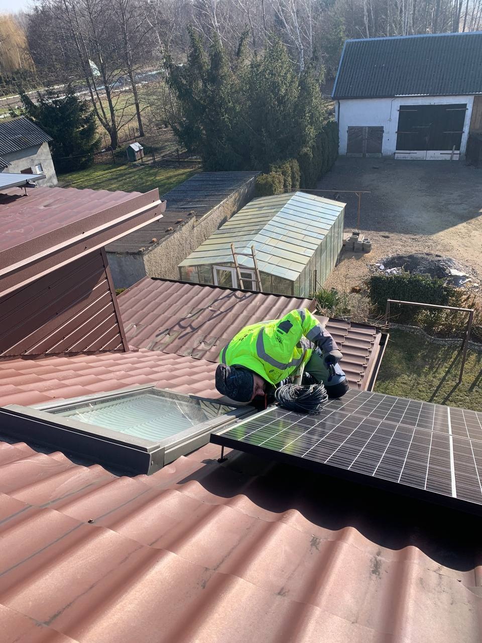 Monter w jaskrawej kamizelce łączy przewody do panelu fotowoltaicznego na dachu z dachówką w kolorze ceglastym, widok z góry na tle zabudowań gospodarczych i szklarni.