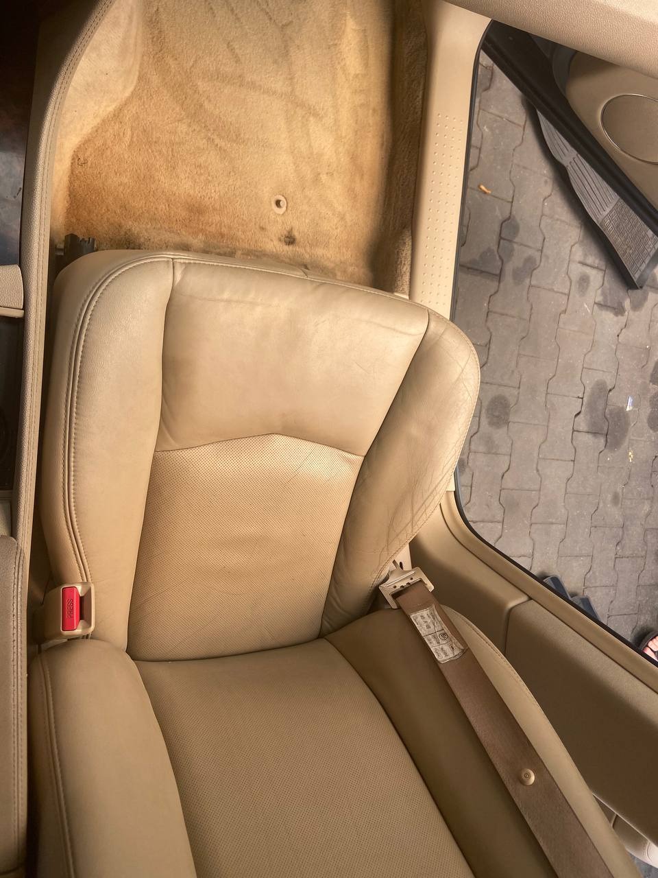 Beżowy skórzany fotel samochodowy z brudną wykładziną podłogową w tle, widoczne zapięcie pasów bezpieczeństwa, wnętrze pojazdu w trakcie demontażu.