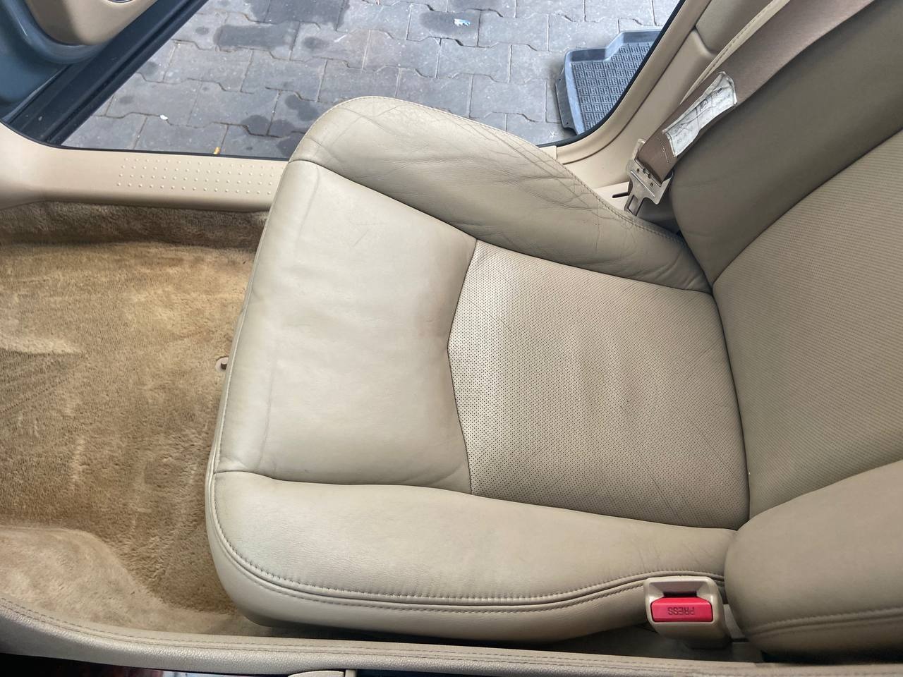 Beżowy skórzany fotel samochodowy z widocznymi śladami użytkowania i plamami, obok beżowa wykładzina podłogowa, widoczny fragment pasa bezpieczeństwa.