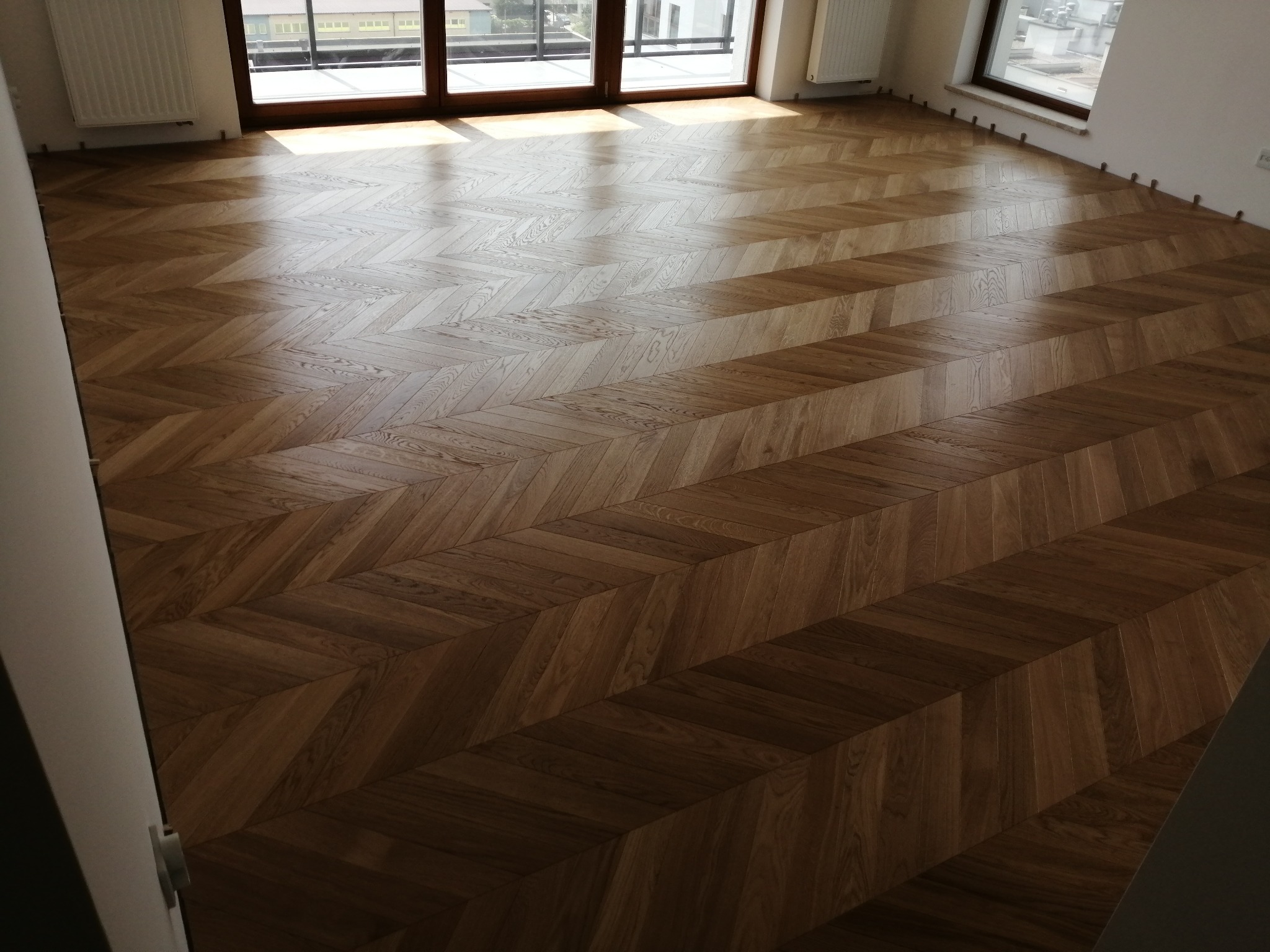 Parkiet ułożony w jodełkę francuską w jasnym pokoju z oknami balkonowymi. Widoczne listwy dystansowe przy ścianach.