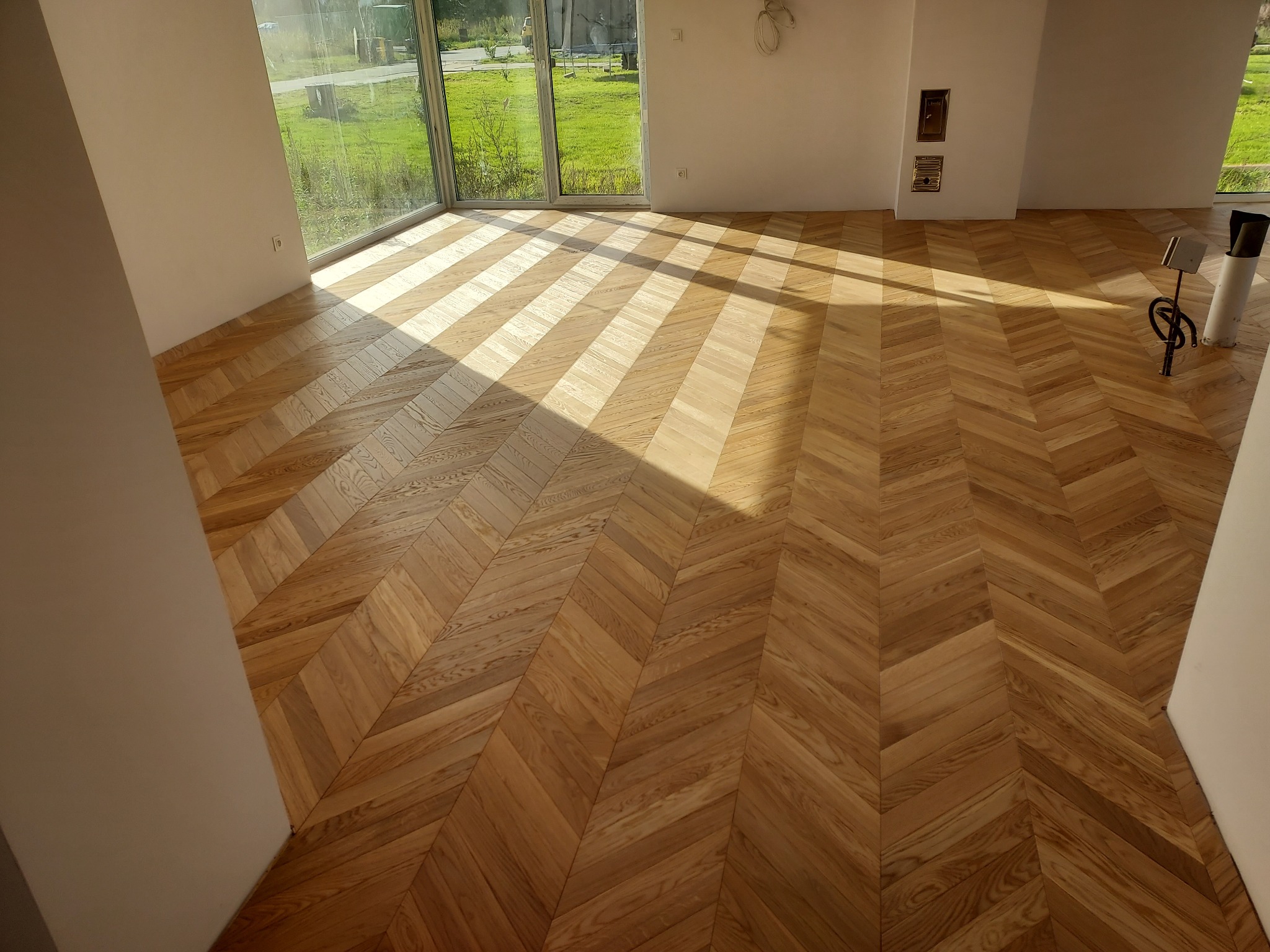 Parkiet ułożony we wzór jodełki w jasnym, przestronnym wnętrzu z dużymi oknami i wpadającym światłem słonecznym. Widoczne detale wykończenia i naturalna kolorystyka drewna.