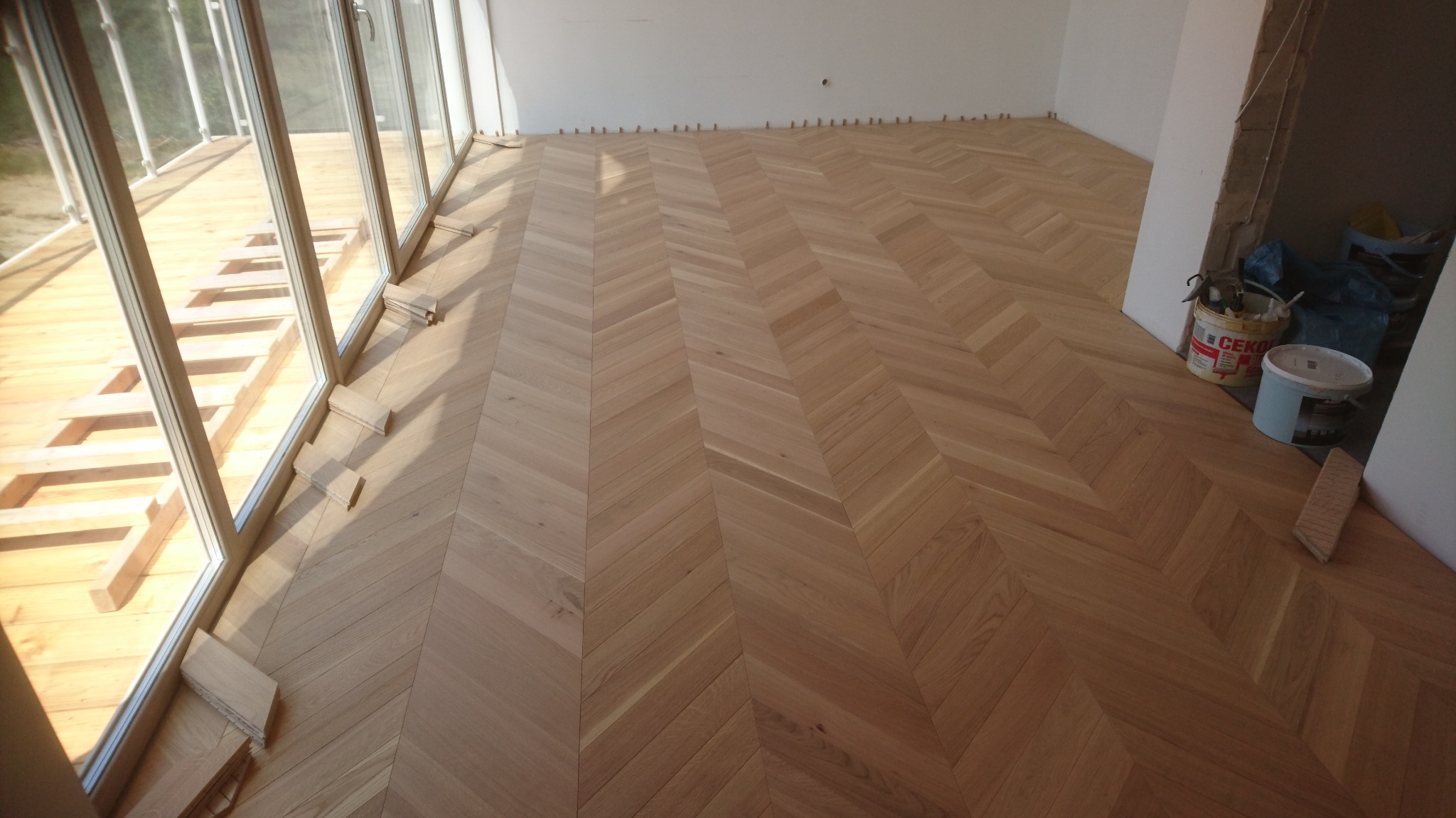 Ułożony parkiet w jodełkę francuską, widoczne kliny dystansowe przy ścianie i duże okno z drewnianą konstrukcją. Wiaderka po kleju w rogu pomieszczenia.