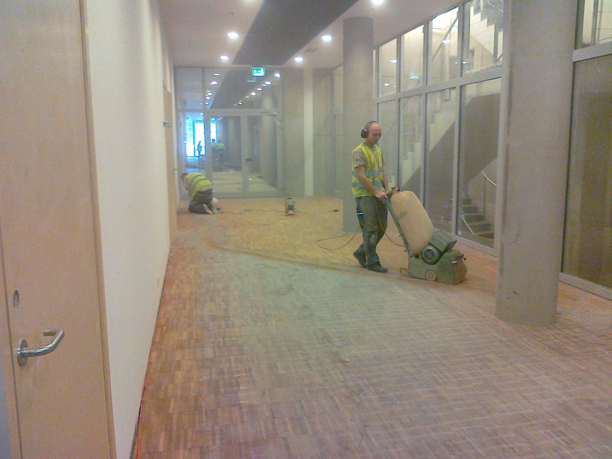 Szlifowanie parkietu w korytarzu biurowym. Pracownik w słuchawkach używa maszyny do cyklinowania, drugi pracuje na kolanach. Widoczny fragment wyszlifowanej podłogi.