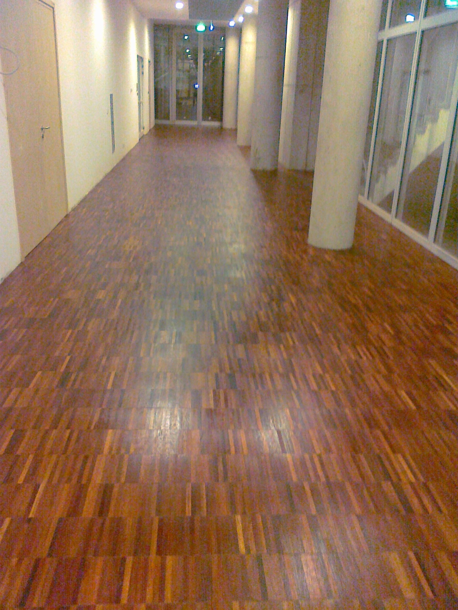 Długi korytarz z parkietem w odcieniach brązu, biegnący wzdłuż białych ścian i kolumn. Widoczne drzwi i duże okna na końcu korytarza.