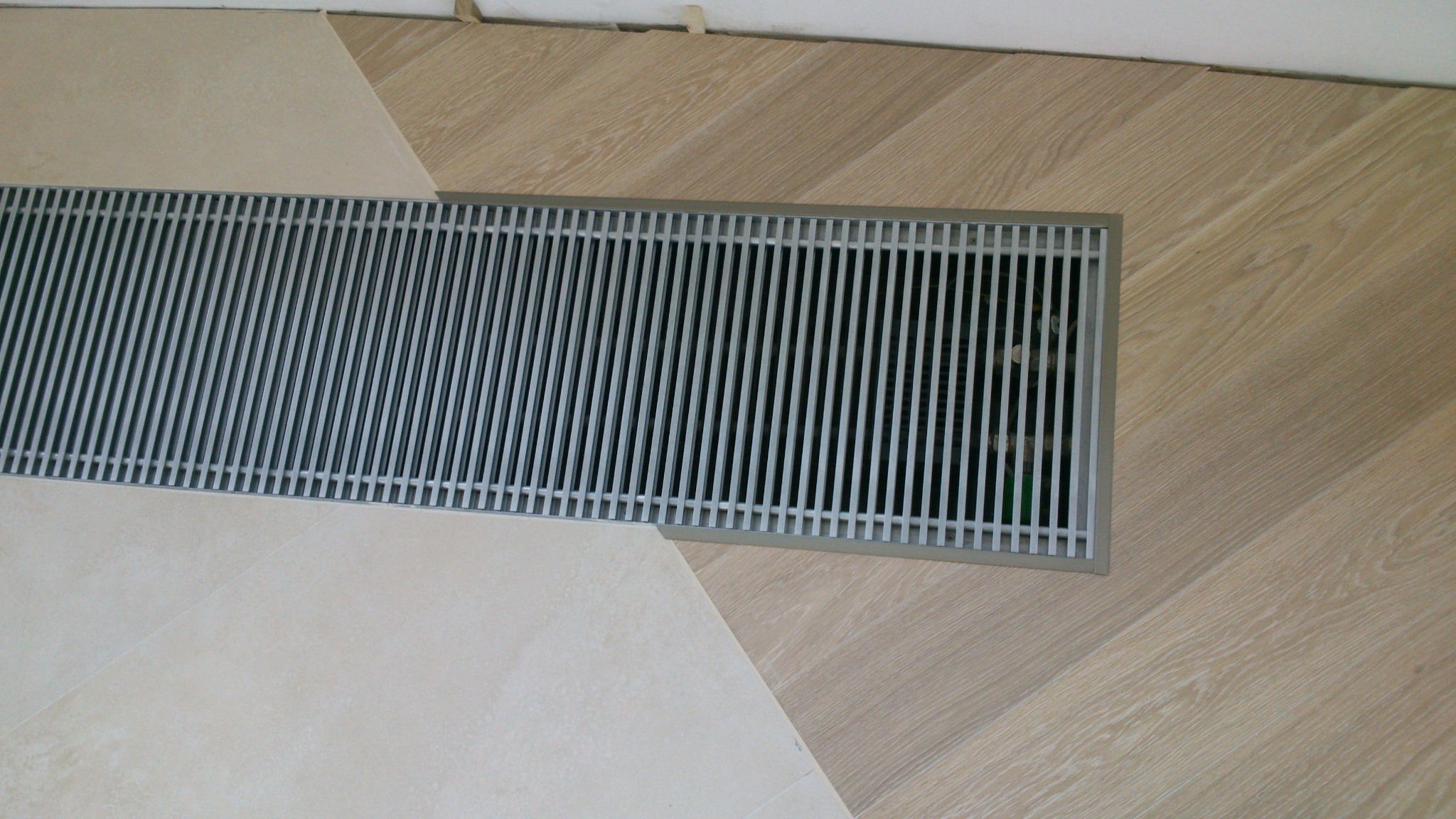 Beżowe panele ułożone w jodełkę, sąsiadujące z jasnymi płytkami i kratką wentylacyjną. Widok z góry na fragment podłogi po remoncie.