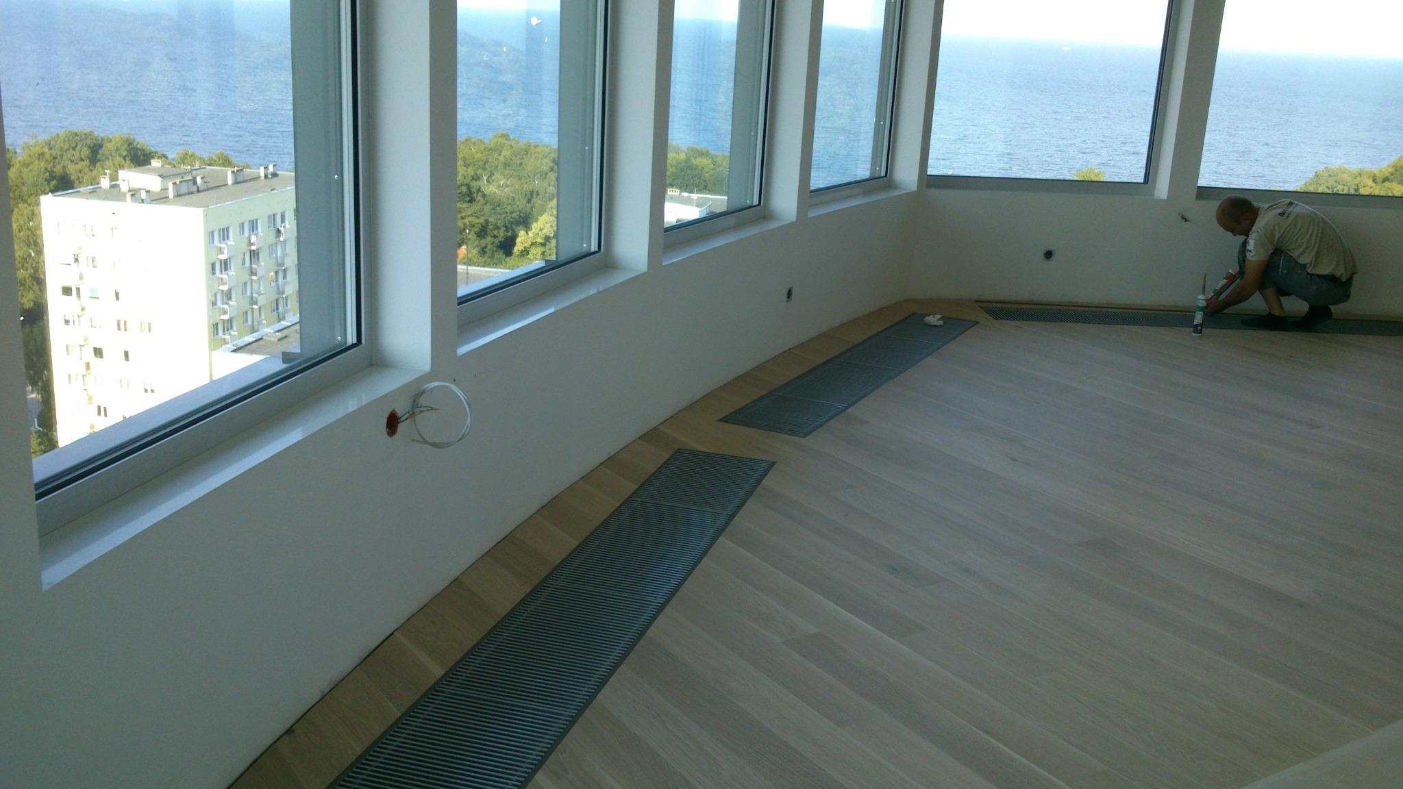 Jasne drewno parkietu ułożonego w jodełkę, z wbudowanymi kratkami wentylacyjnymi. Pracownik aplikuje uszczelniacz przy oknie z widokiem na morze.